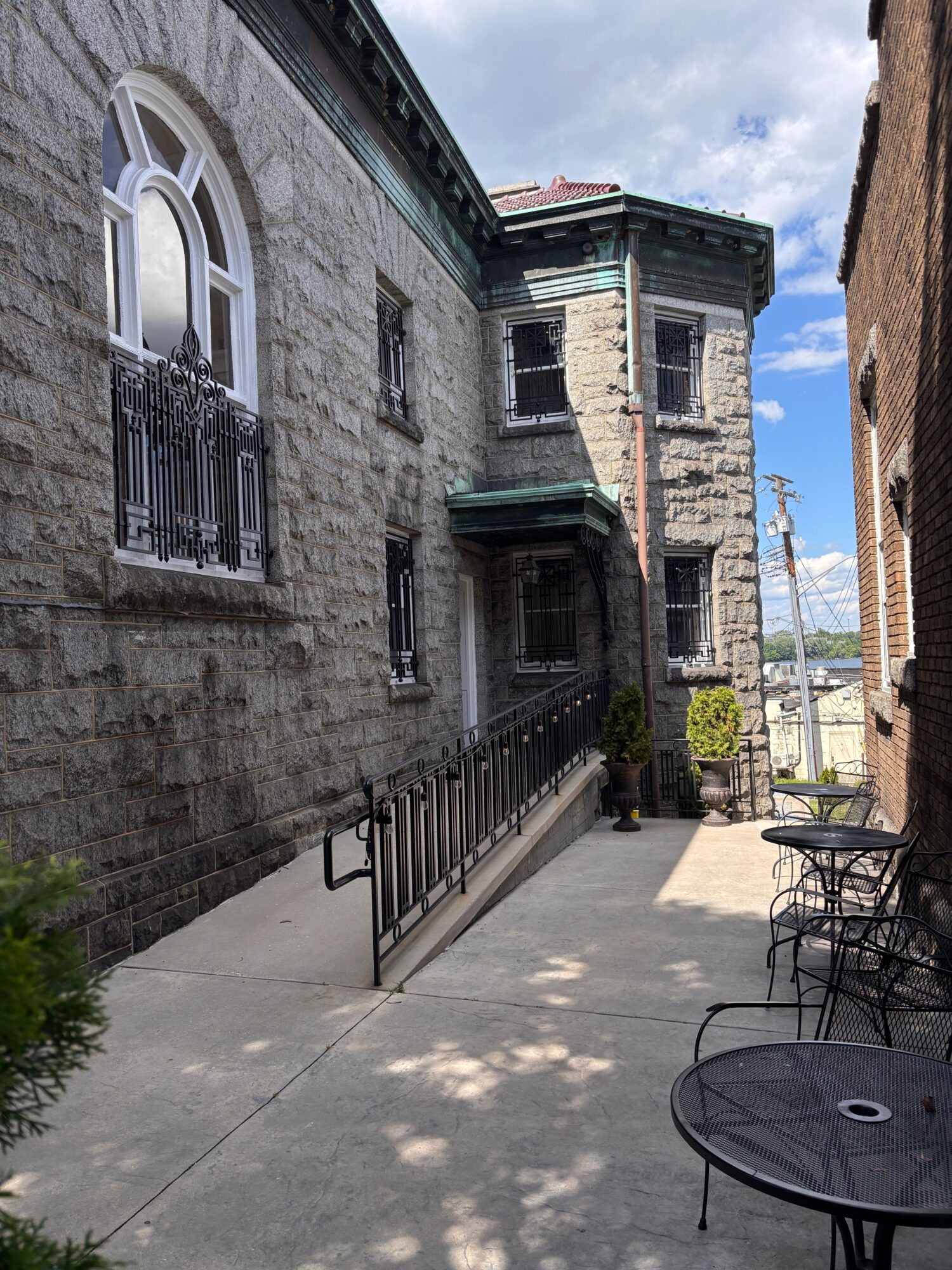 View of a stone building with a window and balcony, outdoor patio with table and chairs, and cityscape in background.