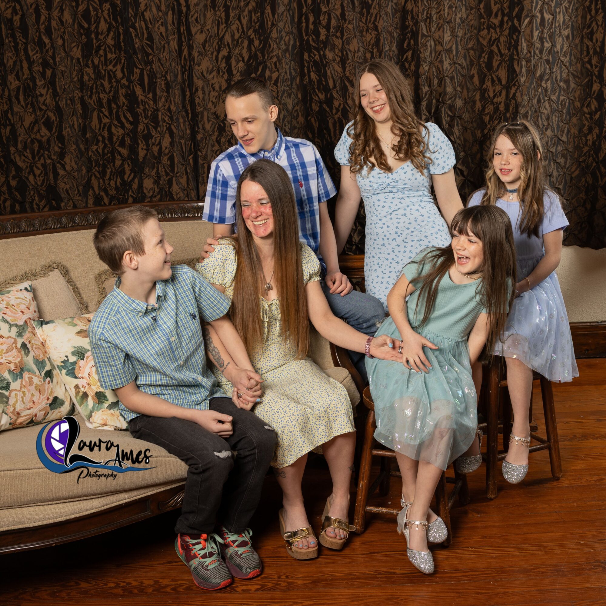 Group of six children and a woman sitting and standing in a room with a dark curtain background, smiling and interacting.