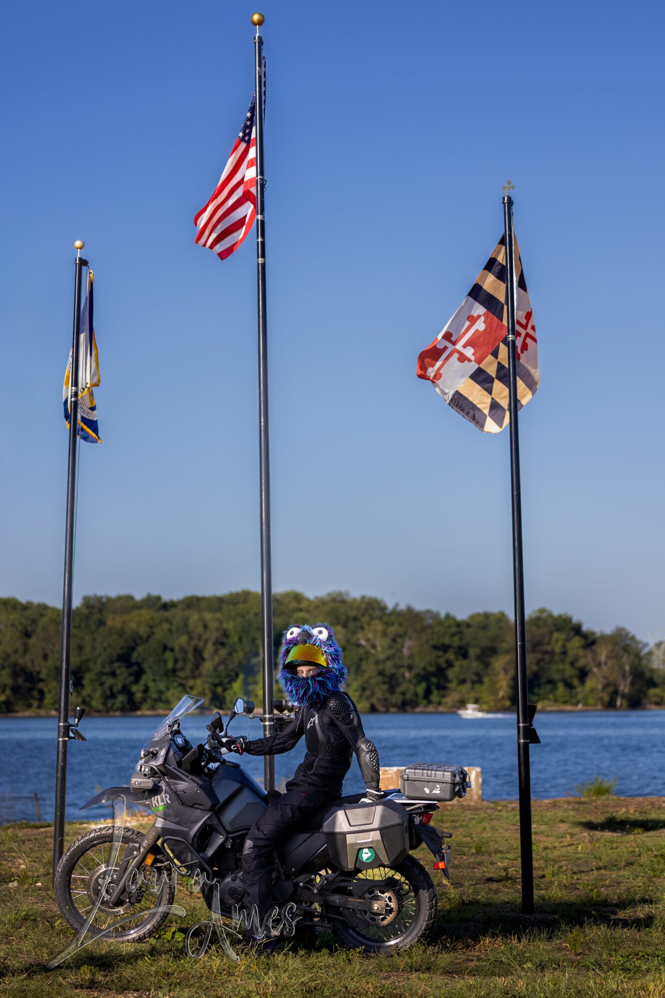 Person in motorcycle gear sitting on a motorcycle near three flagpoles with flags, water, and trees in background.