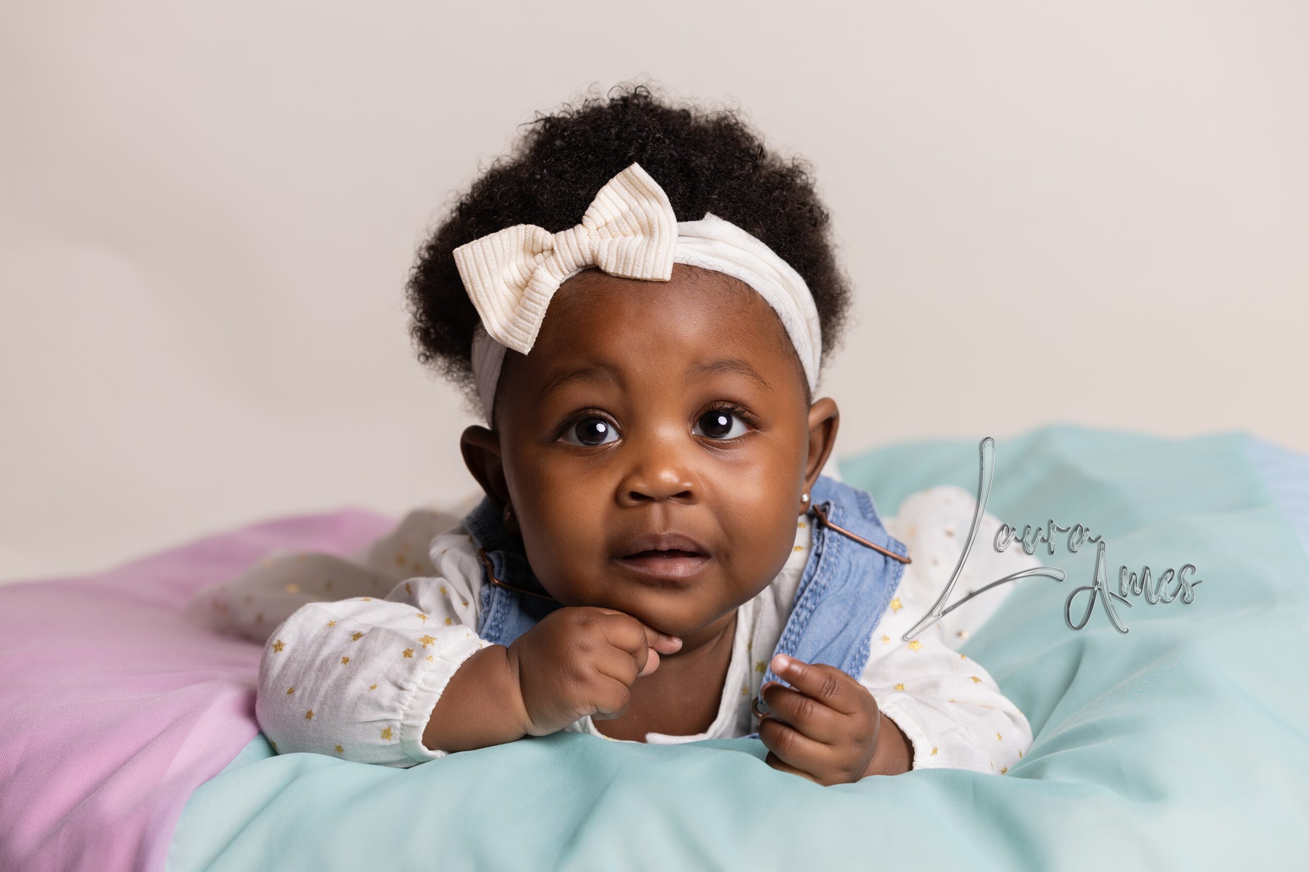 Young girl with a white bow headband lying on her stomach on a bed, looking at the camera.