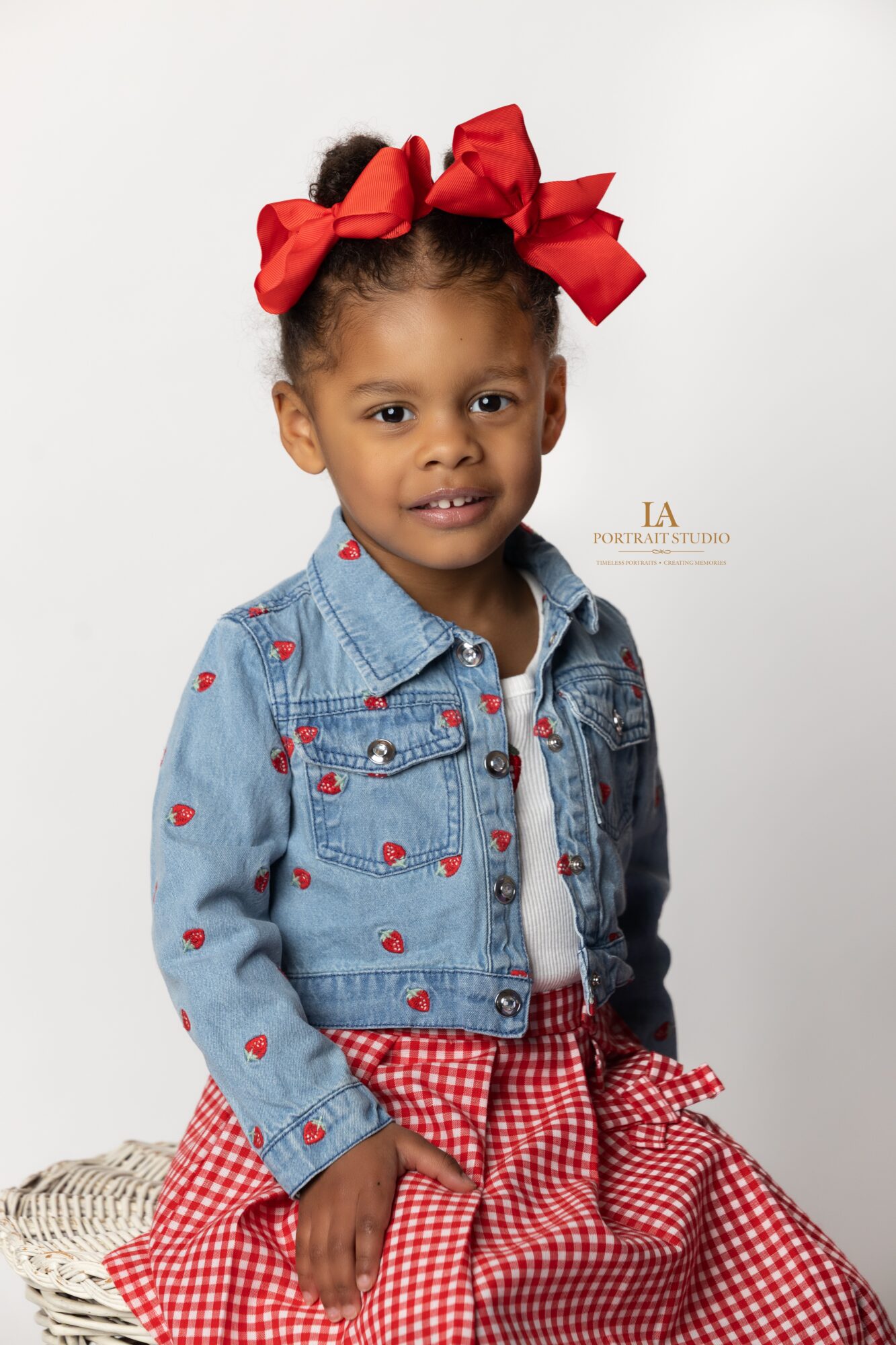 Young girl with two large red bows in her hair, wearing a denim jacket and red checkered skirt, smiling.