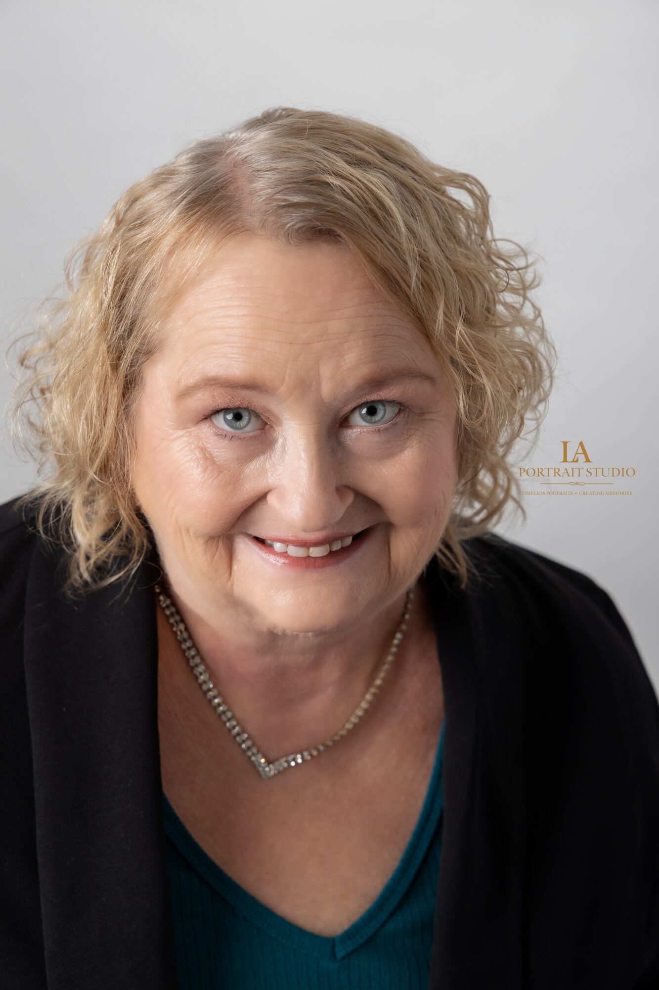 Portrait of a woman with curly blonde hair smiling, wearing a black jacket and a necklace, against a plain background.