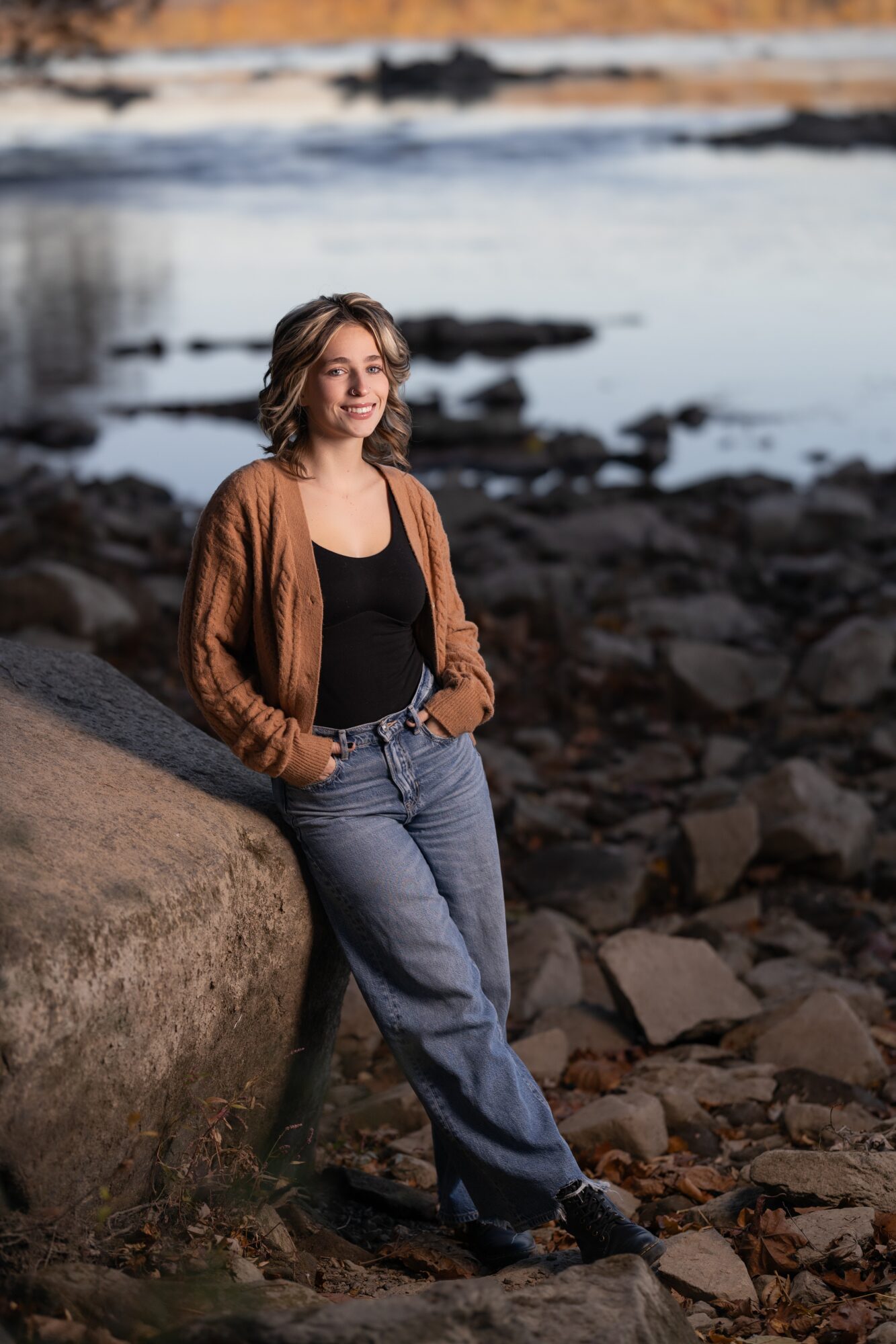 Woman standing outdoors near rocks and water, wearing a brown cardigan and jeans, looking to the side.