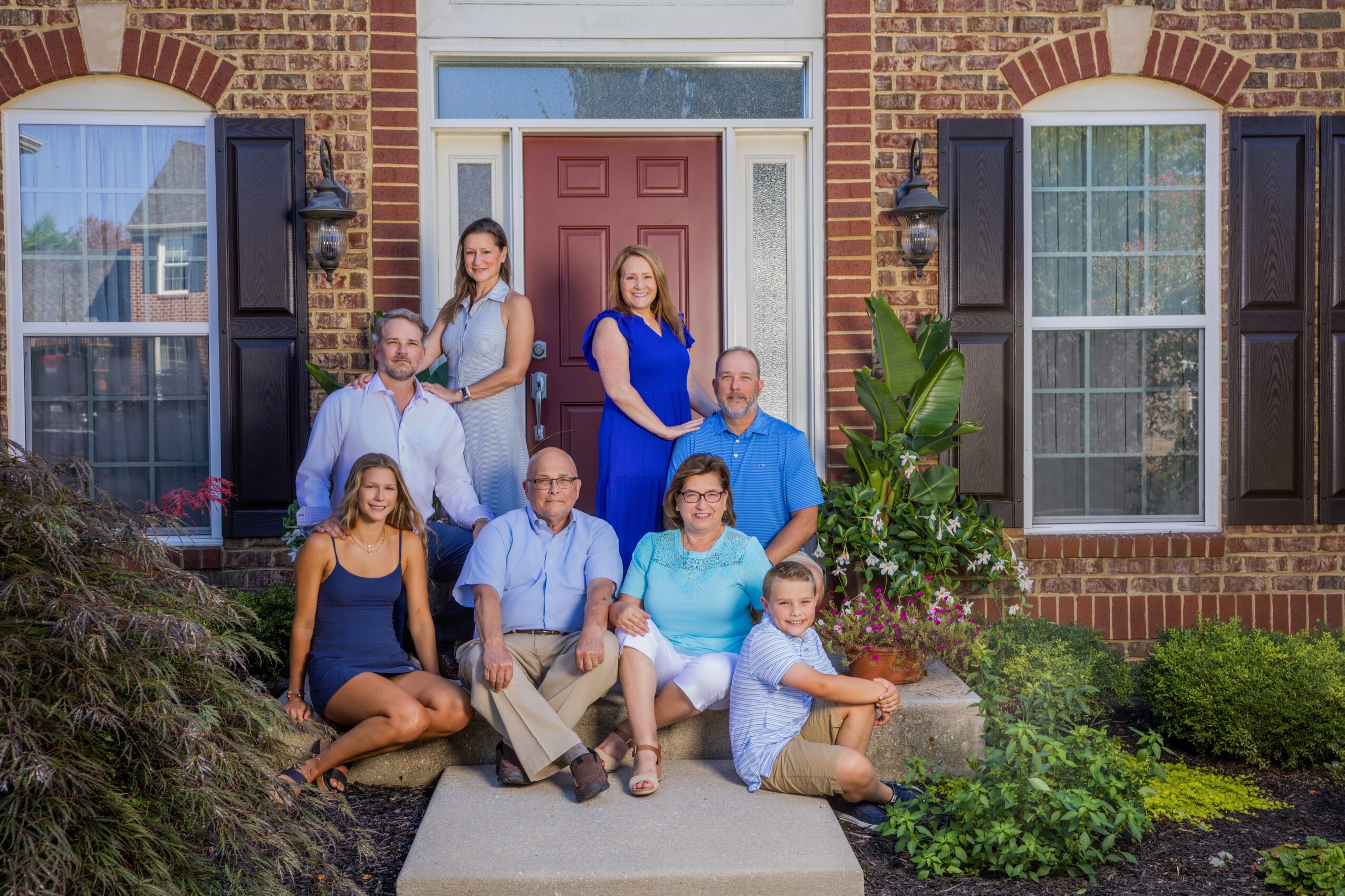 Family of nine posing outside a brick house with black shutters, some sitting and others standing, smiling.