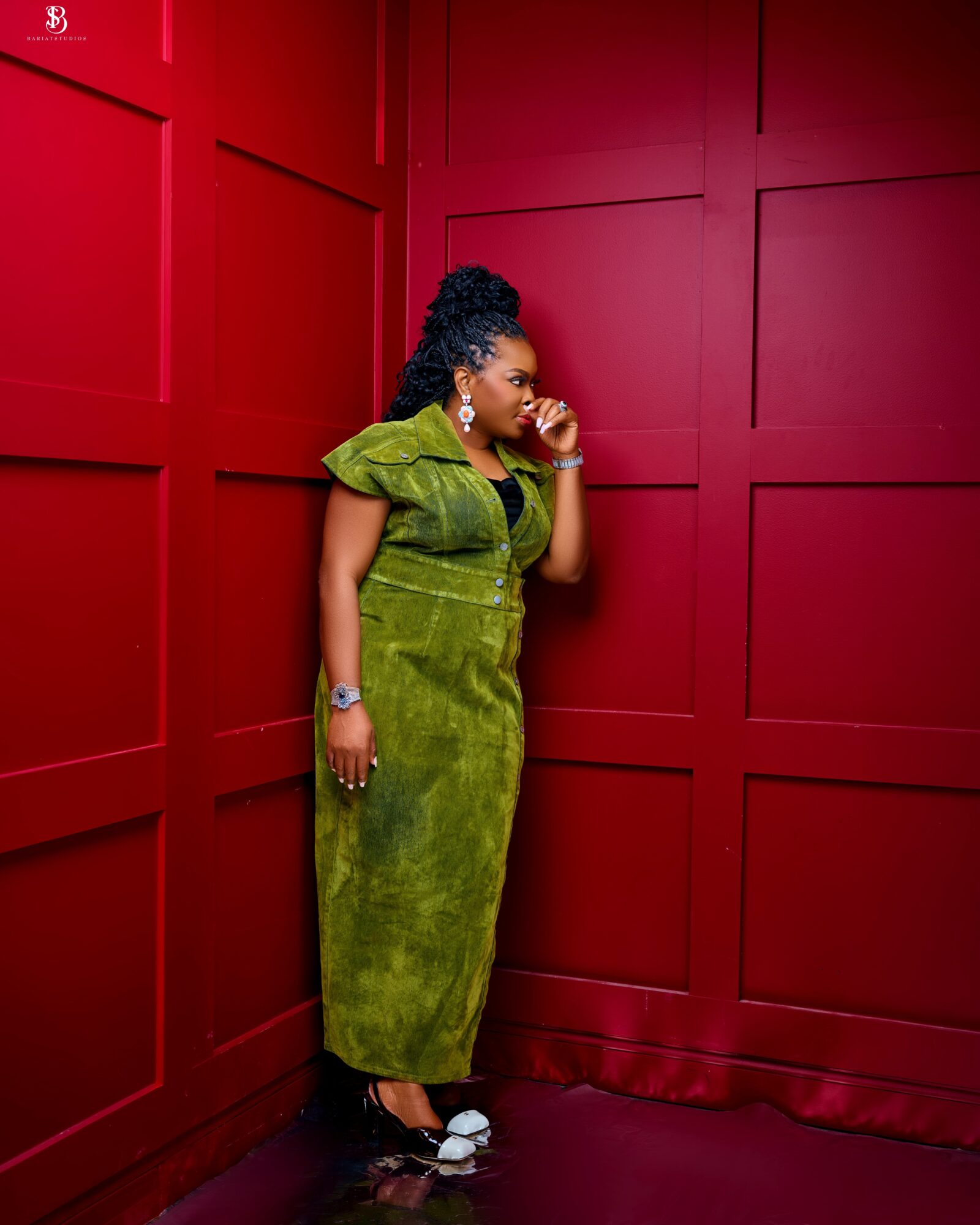 Woman in green dress standing against red paneled wall, talking on phone, wearing jewelry and white shoes.