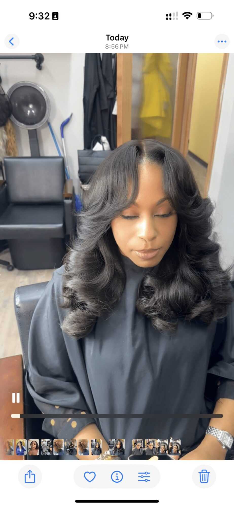 Woman with styled hair sitting in a salon chair, wearing a black cape, with salon equipment and clothing in the background.