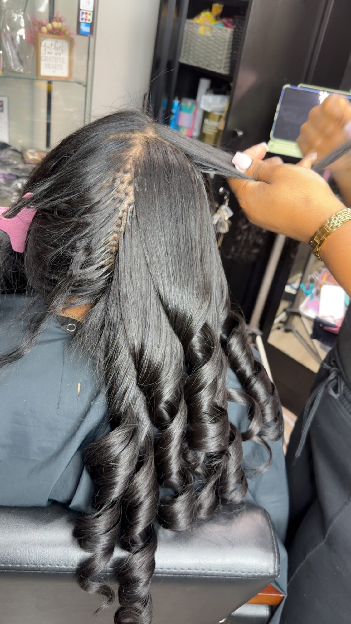 Person with long, black, curled hair sitting in a salon chair, with a stylist taking a photo of their hair.
