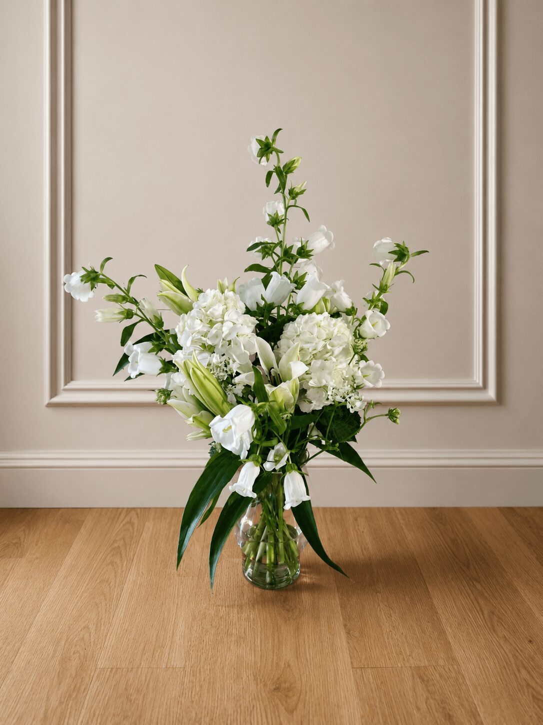 White flower bouquet in a glass vase on a wooden floor against a beige wall with decorative molding