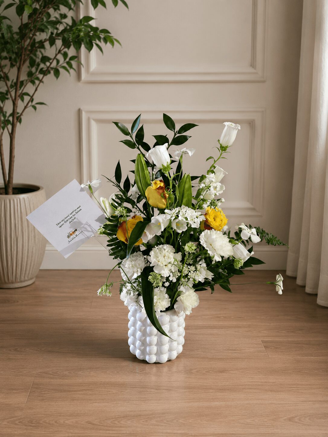 Flower arrangement with white, yellow, and green flowers in a textured white vase on a wooden table, with a potted plant nearby.