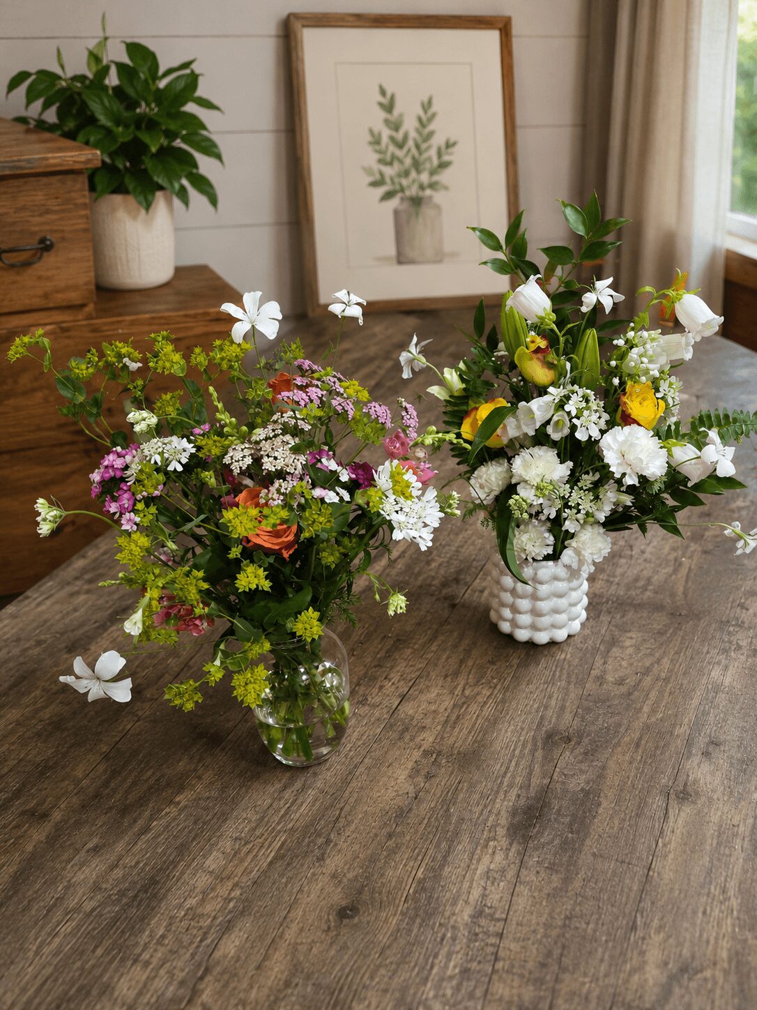 Two floral arrangements on a wooden table with a framed botanical print, potted plant, and window in background.