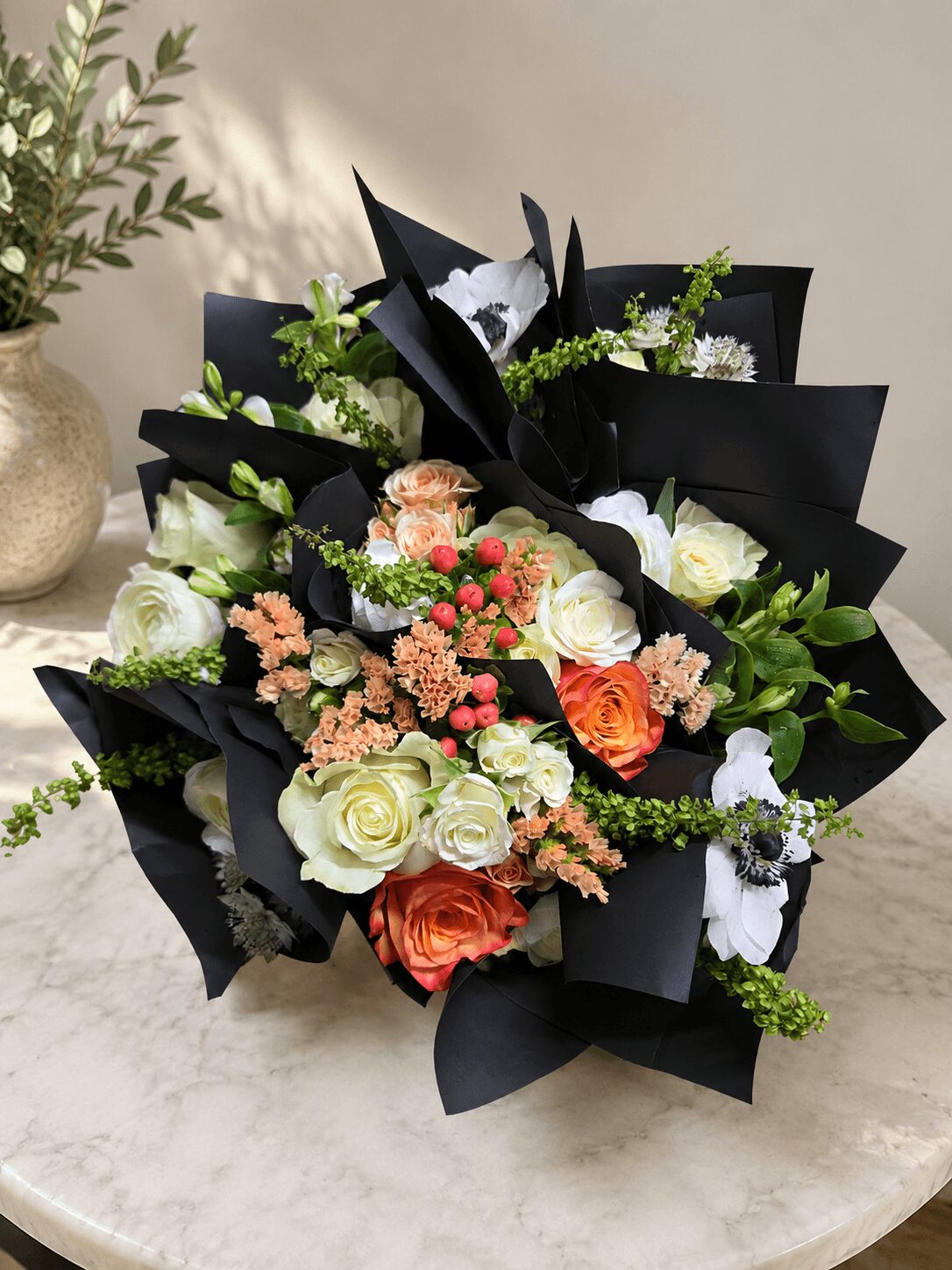 Bouquet of white, peach, and orange flowers with green foliage, wrapped in black paper, on a marble surface.