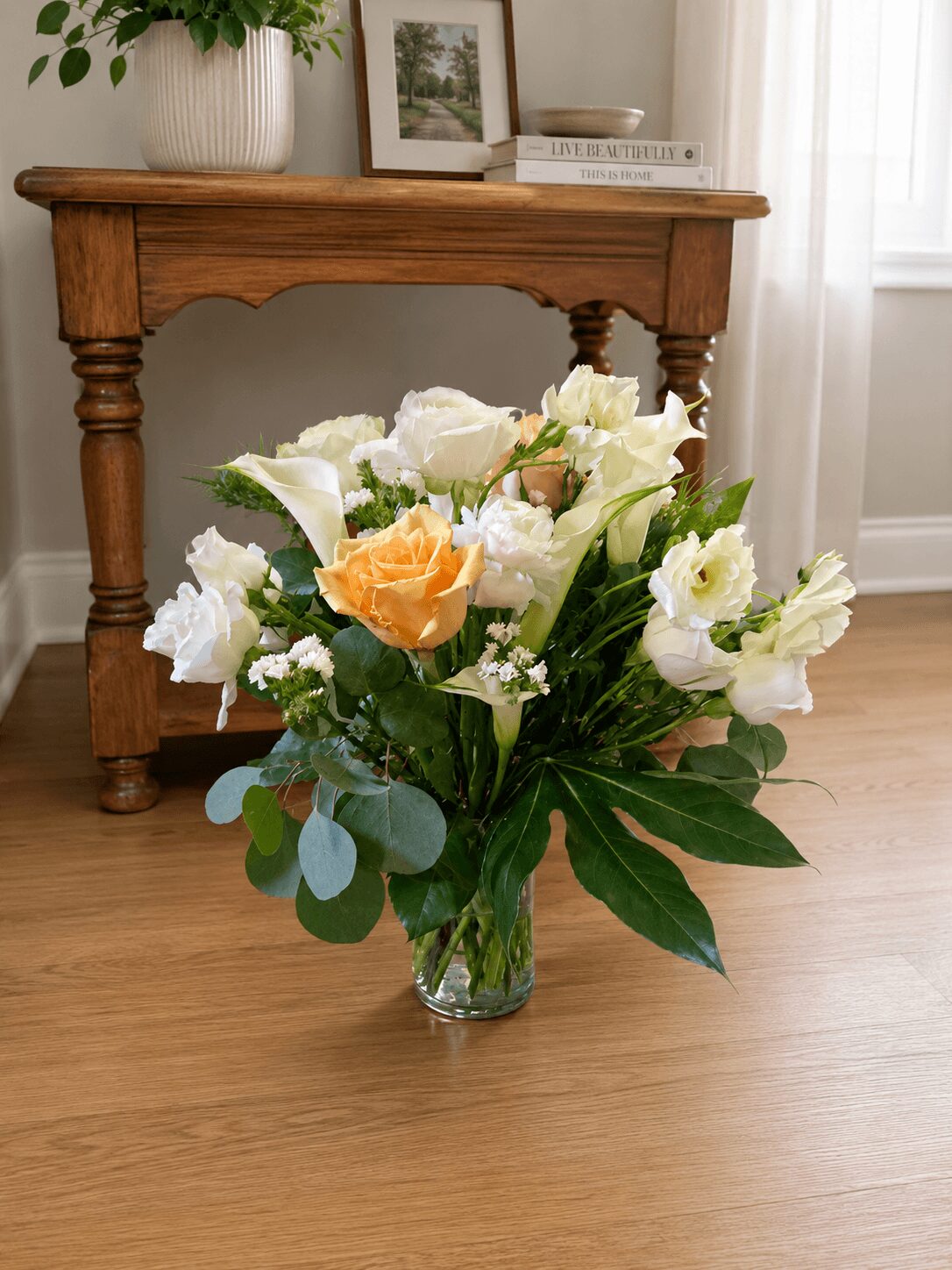 Glass vase with white and peach flowers on a wooden floor, with a wooden table and framed pictures in the background.