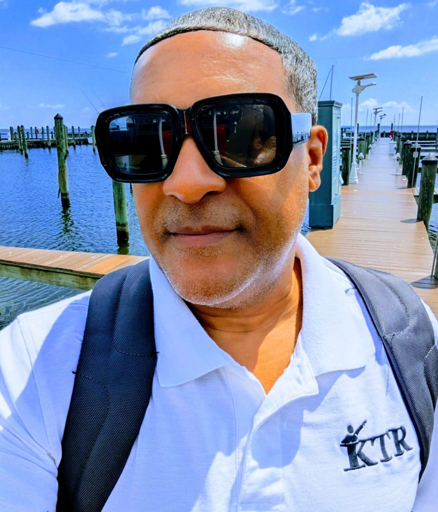 Man with sunglasses and backpack standing on a dock by water, blue sky with clouds in background.