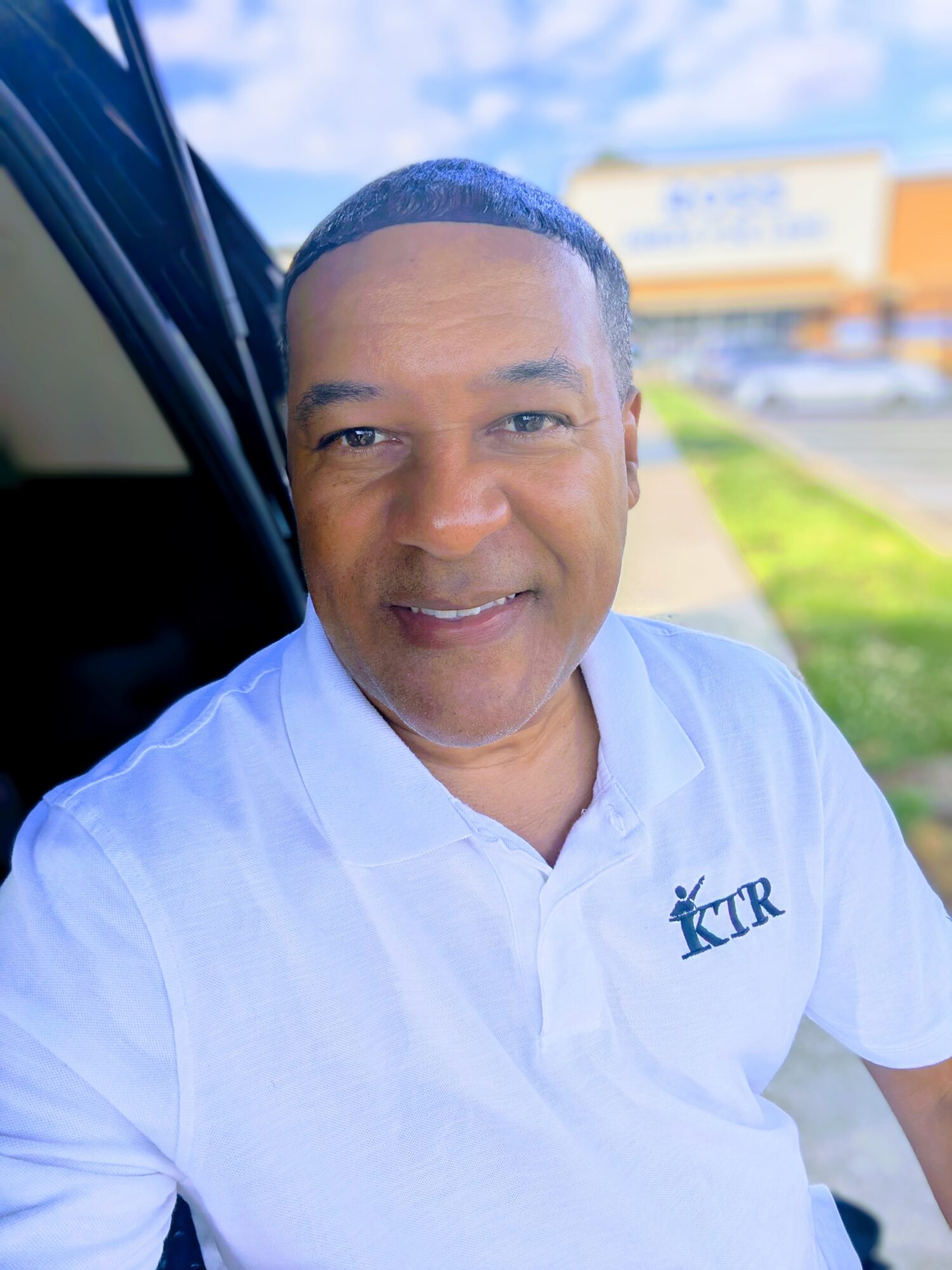 Man in white polo shirt outdoors, smiling, with a blurred background of a building and sky.