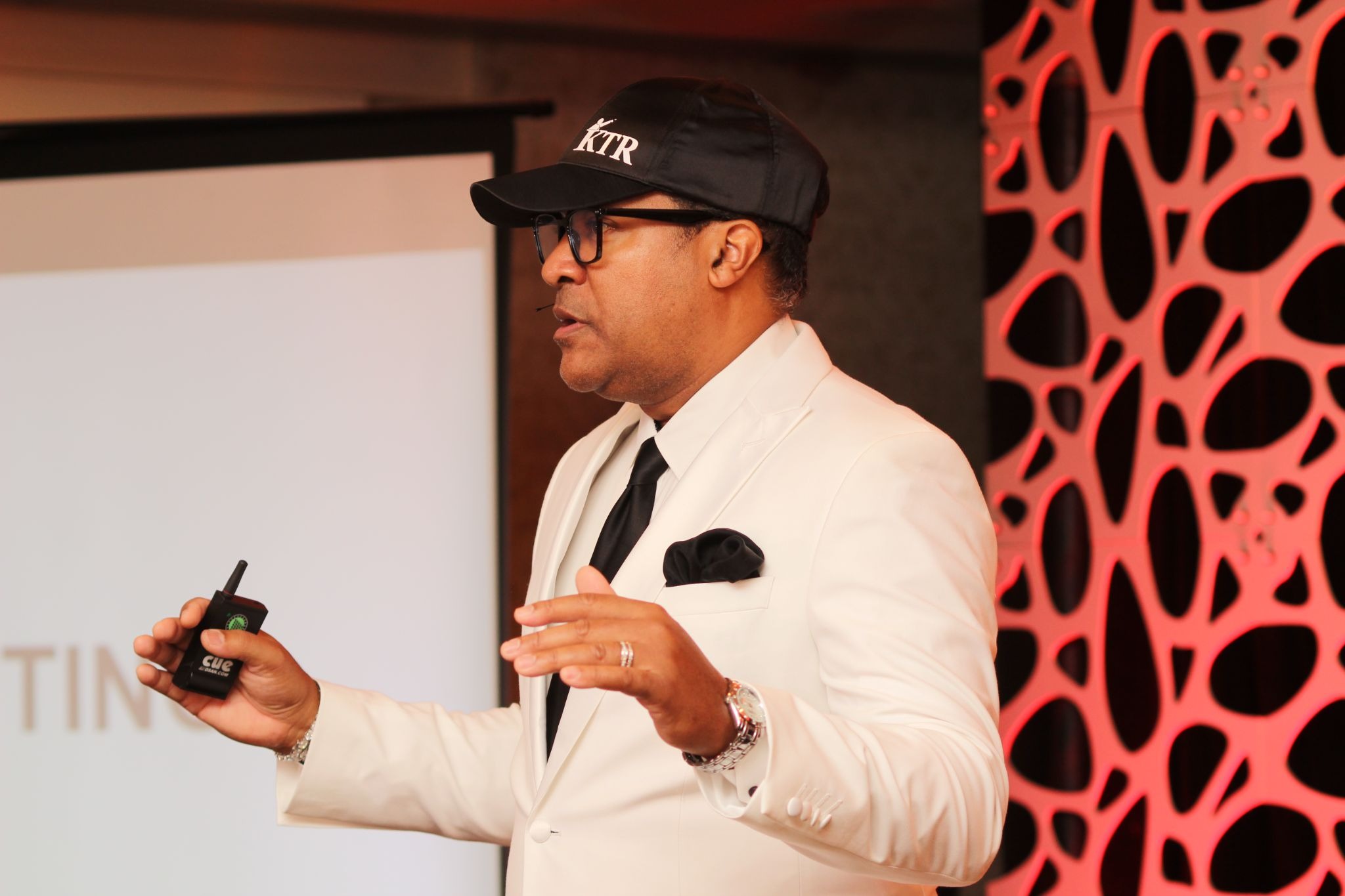 Man in white suit and black cap speaking at a presentation, holding a remote, with a patterned red and black background.