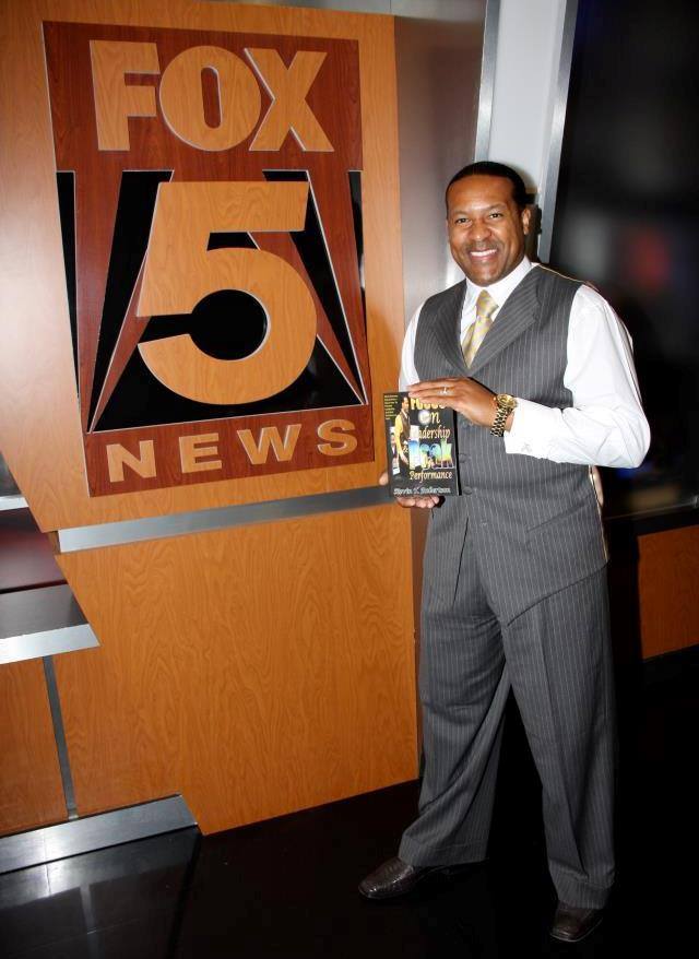 Man in suit holding a tablet in front of a FOX 5 News sign, smiling, in a studio setting.
