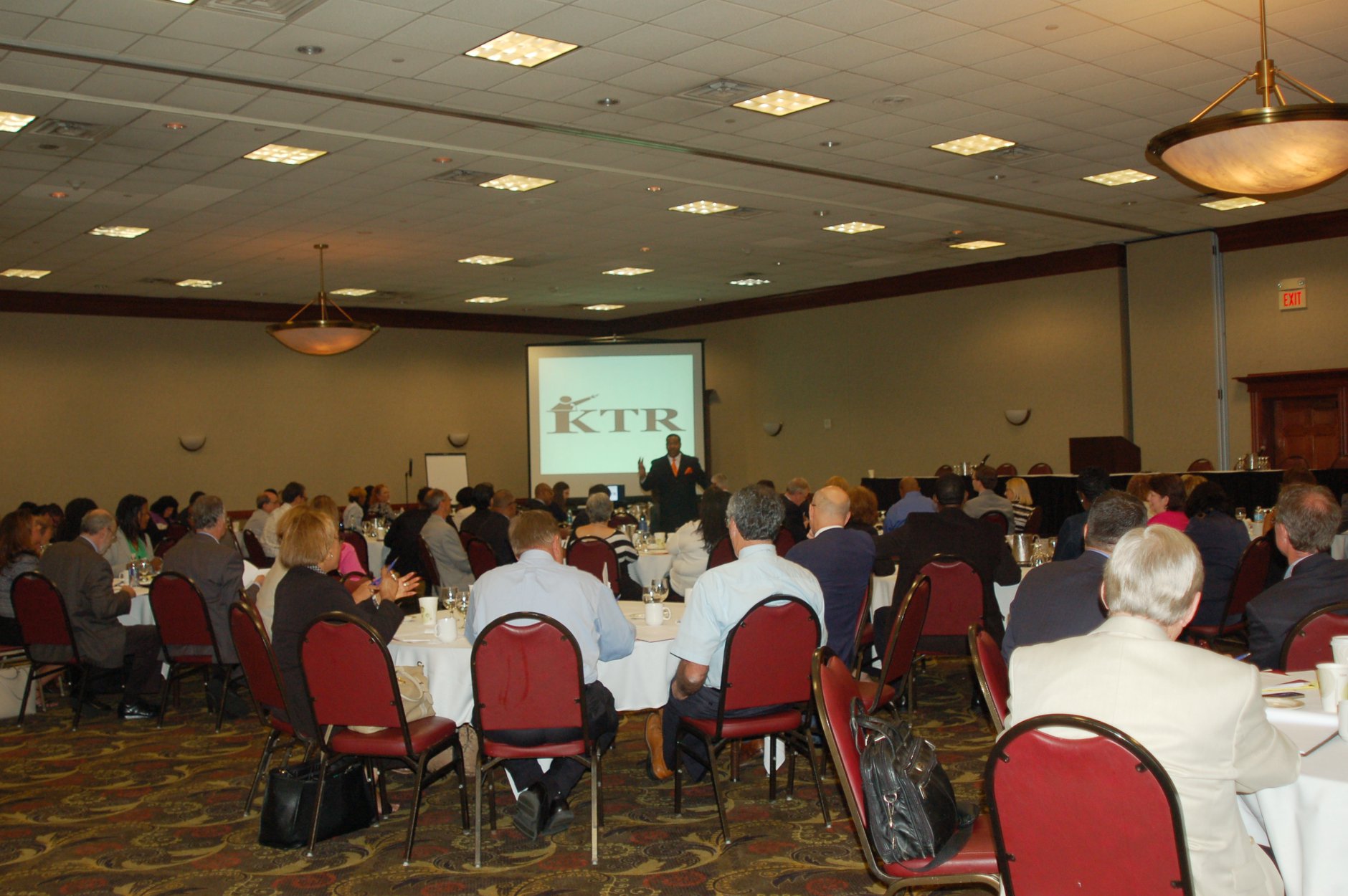 Conference room filled with seated attendees facing a stage with a presentation screen displaying a logo, carpeted floor, and ceiling lights.