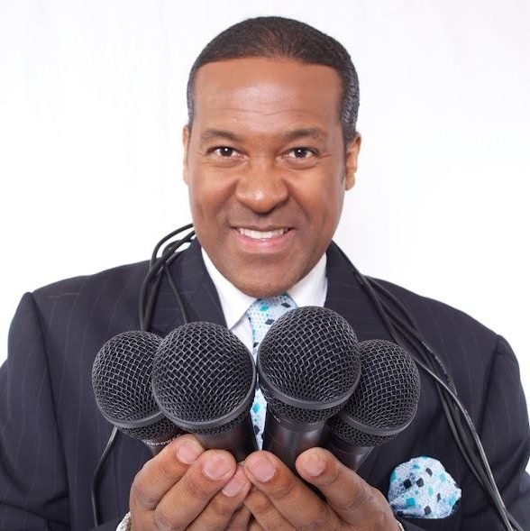 Man in suit holding five microphones, smiling, against a white background.