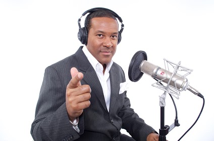 Man in suit wearing headphones, pointing at camera, speaking into a microphone, white background.