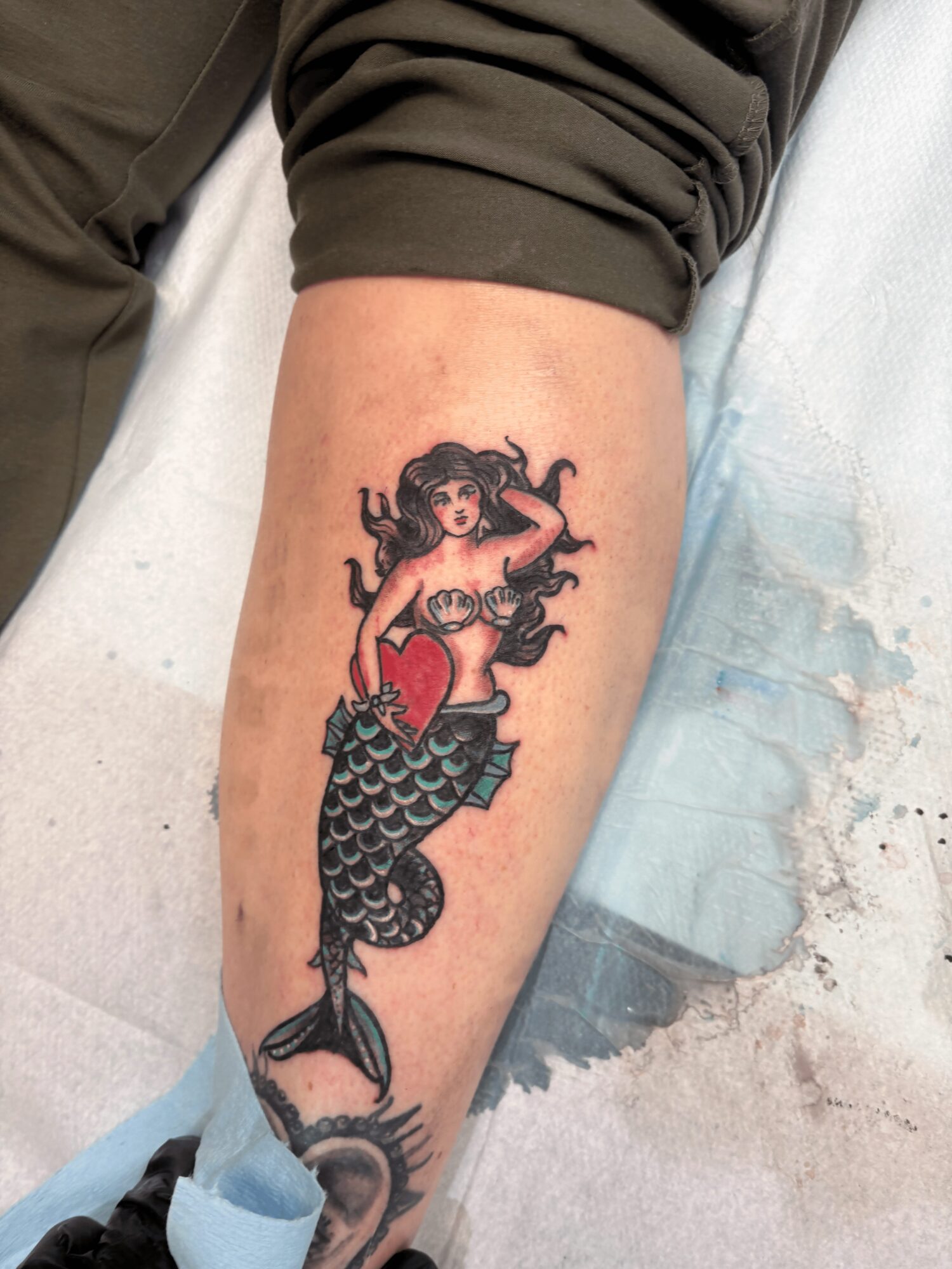 Tattoo of a mermaid with dark hair, holding a red heart, with a fish tail, on person's calf.