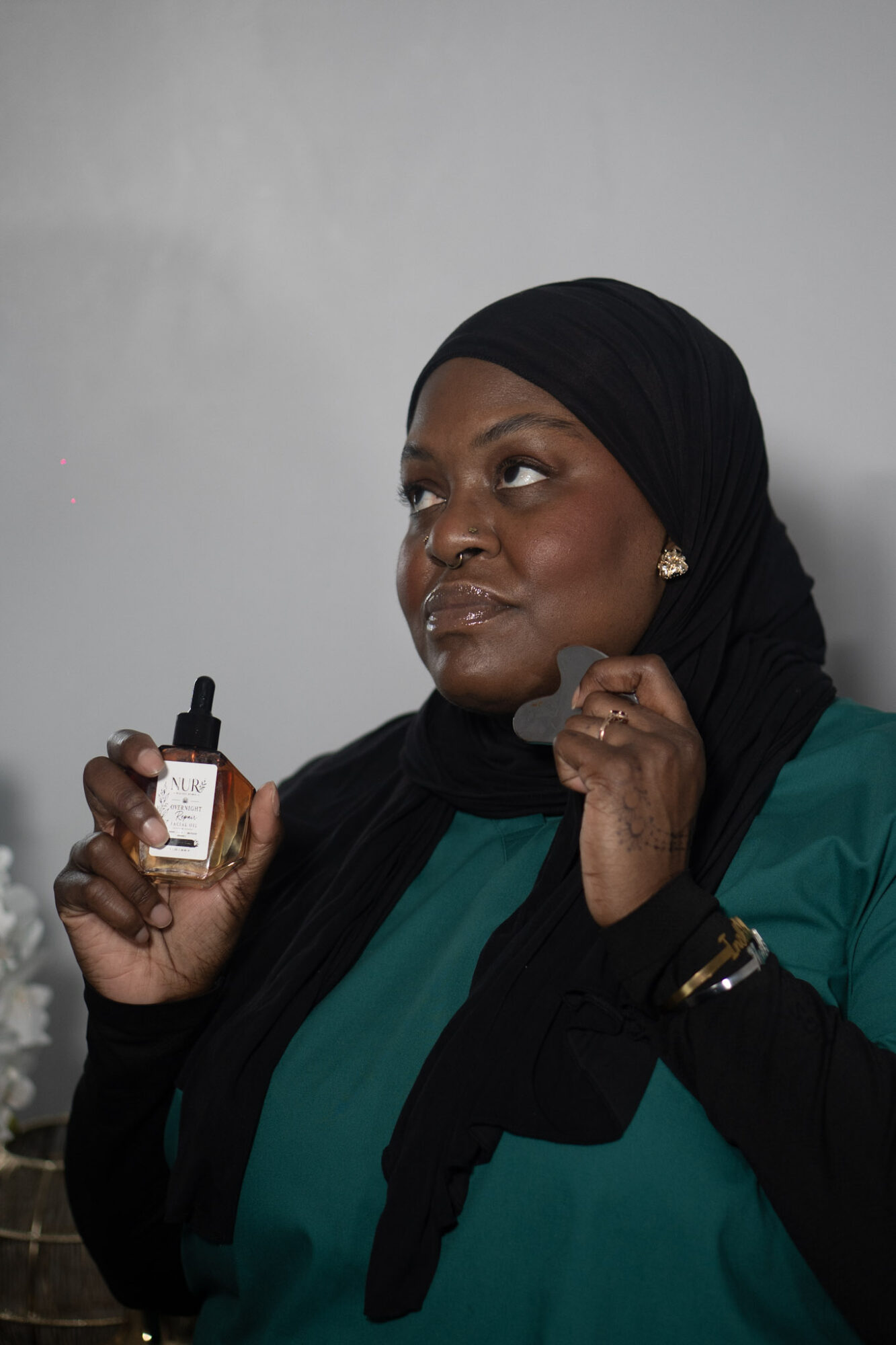 Woman wearing a black headscarf and teal top holding a small bottle, looking thoughtfully to the side.