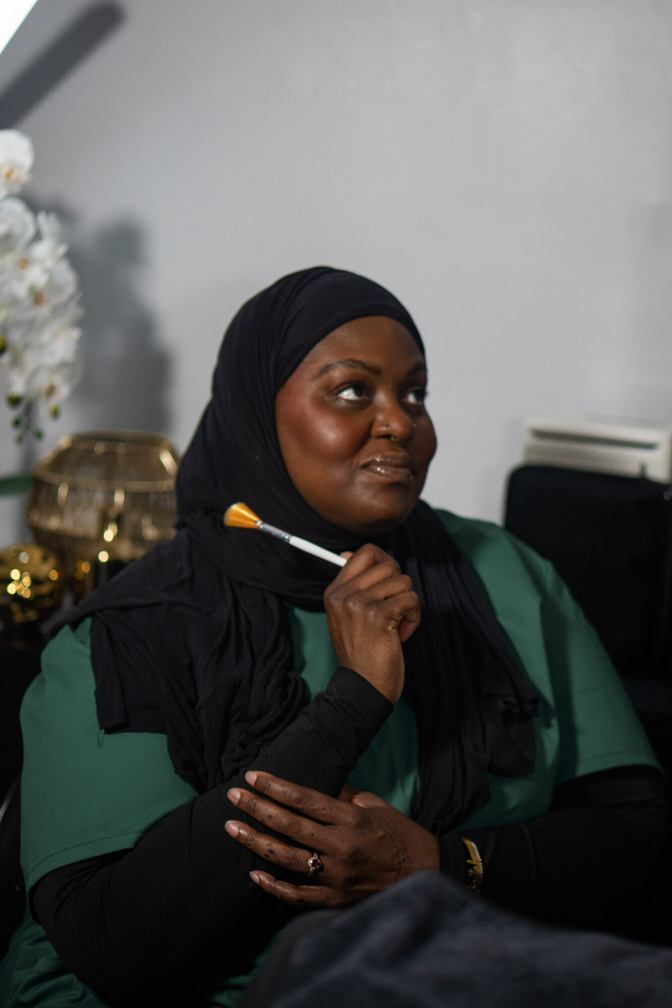 Woman wearing a black headscarf and green top, holding a paintbrush near her face, sitting indoors with a white wall background.