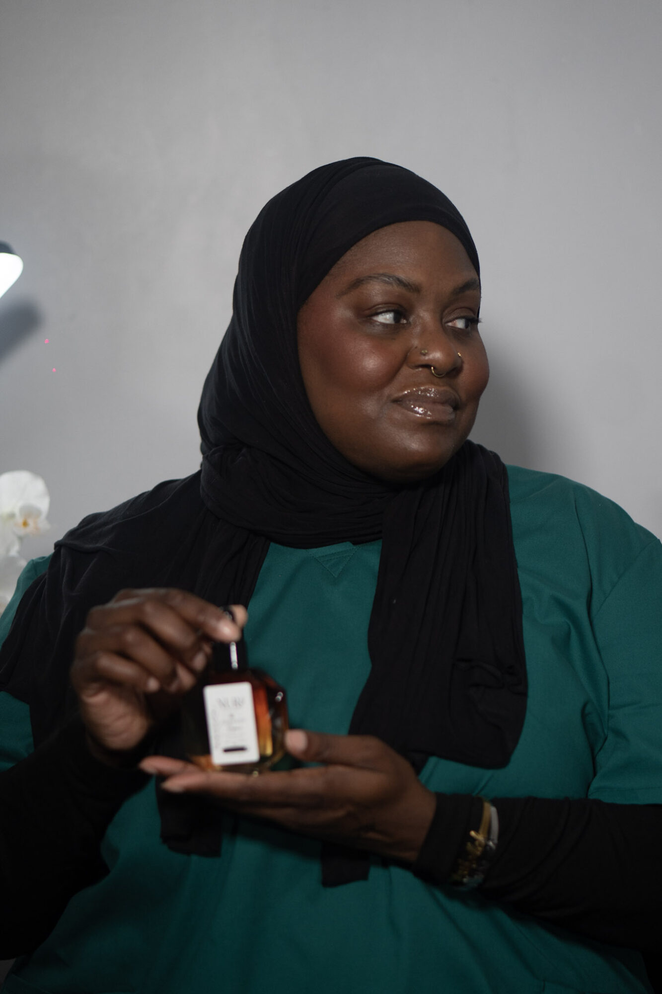 Woman wearing a black headscarf and teal top holding a small device, smiling slightly, against a plain background.