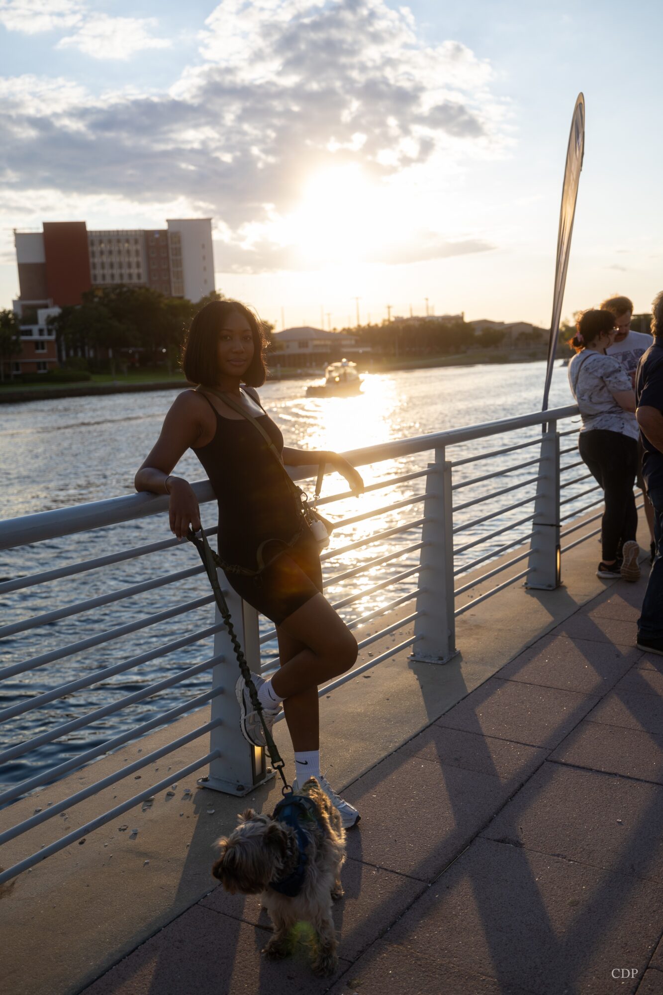 Woman standing by a railing with a dog, overlooking a river at sunset, with people in the background.