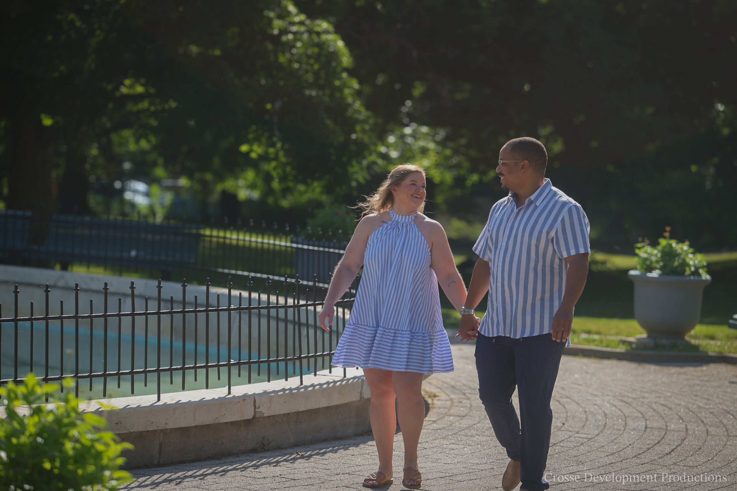 A couple holding hands walking outdoors near a pool, with trees and greenery in the background.