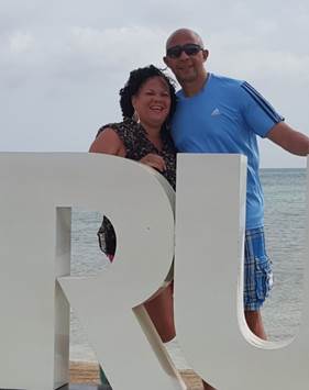 Two people smiling at the beach, standing behind a large white letter R, with ocean and cloudy sky in background.