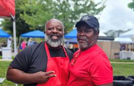 Two men smiling outdoors, one wearing a black shirt and red apron, the other in a red shirt and black cap.