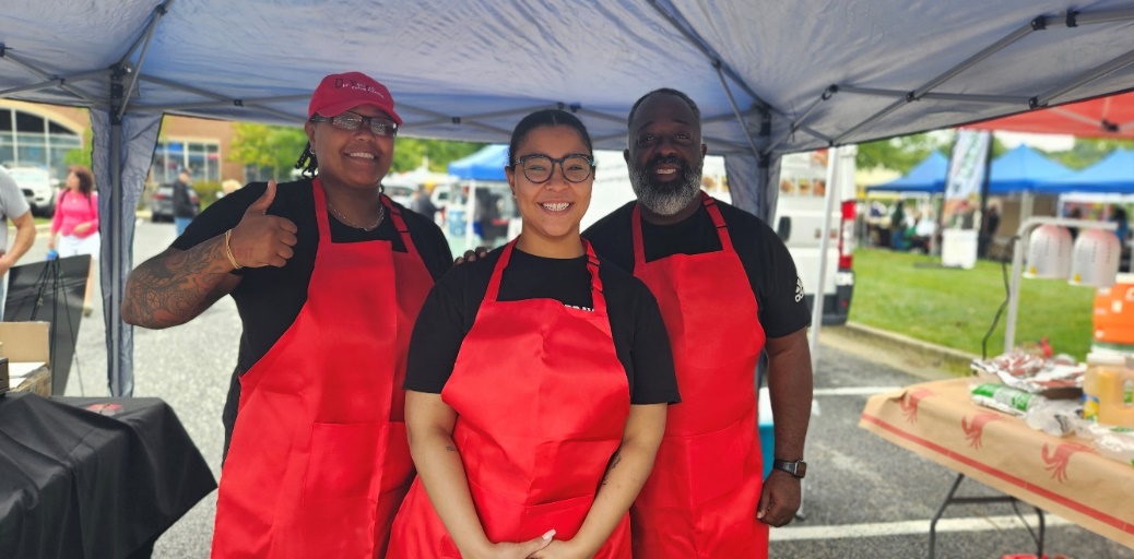 Three people wearing red aprons and black shirts standing under a tent outdoors, smiling at the camera.