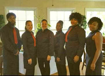 Six people standing indoors in front of windows, dressed in dark clothing with red accents, smiling.