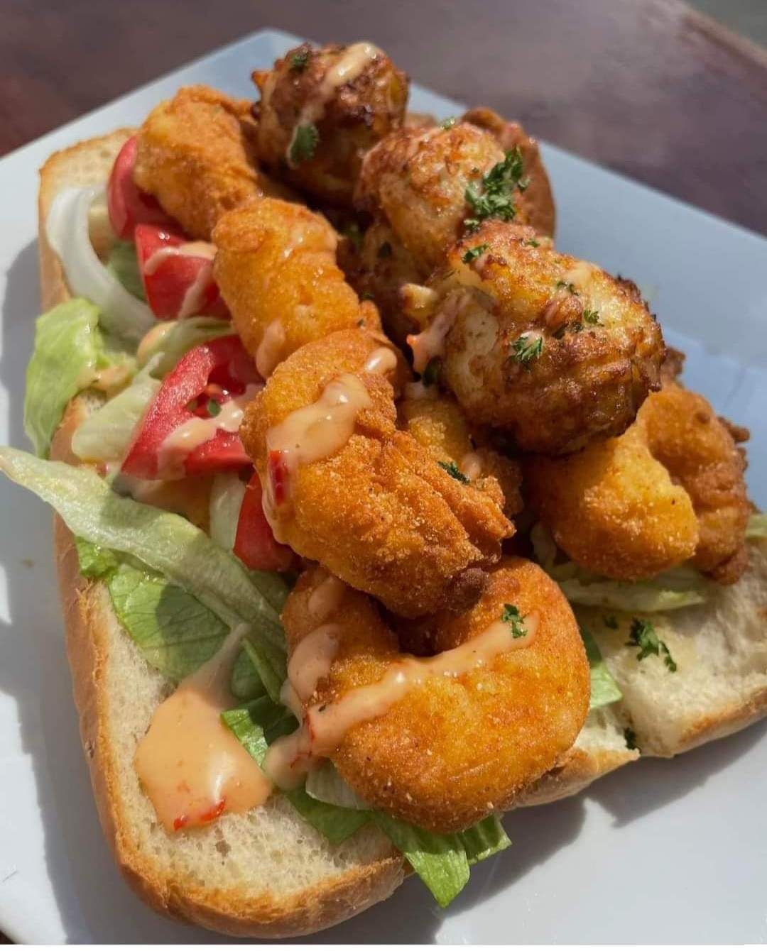 Fried shrimp and meatballs on a sandwich with lettuce, tomato, and sauce on bread, served on a white plate.