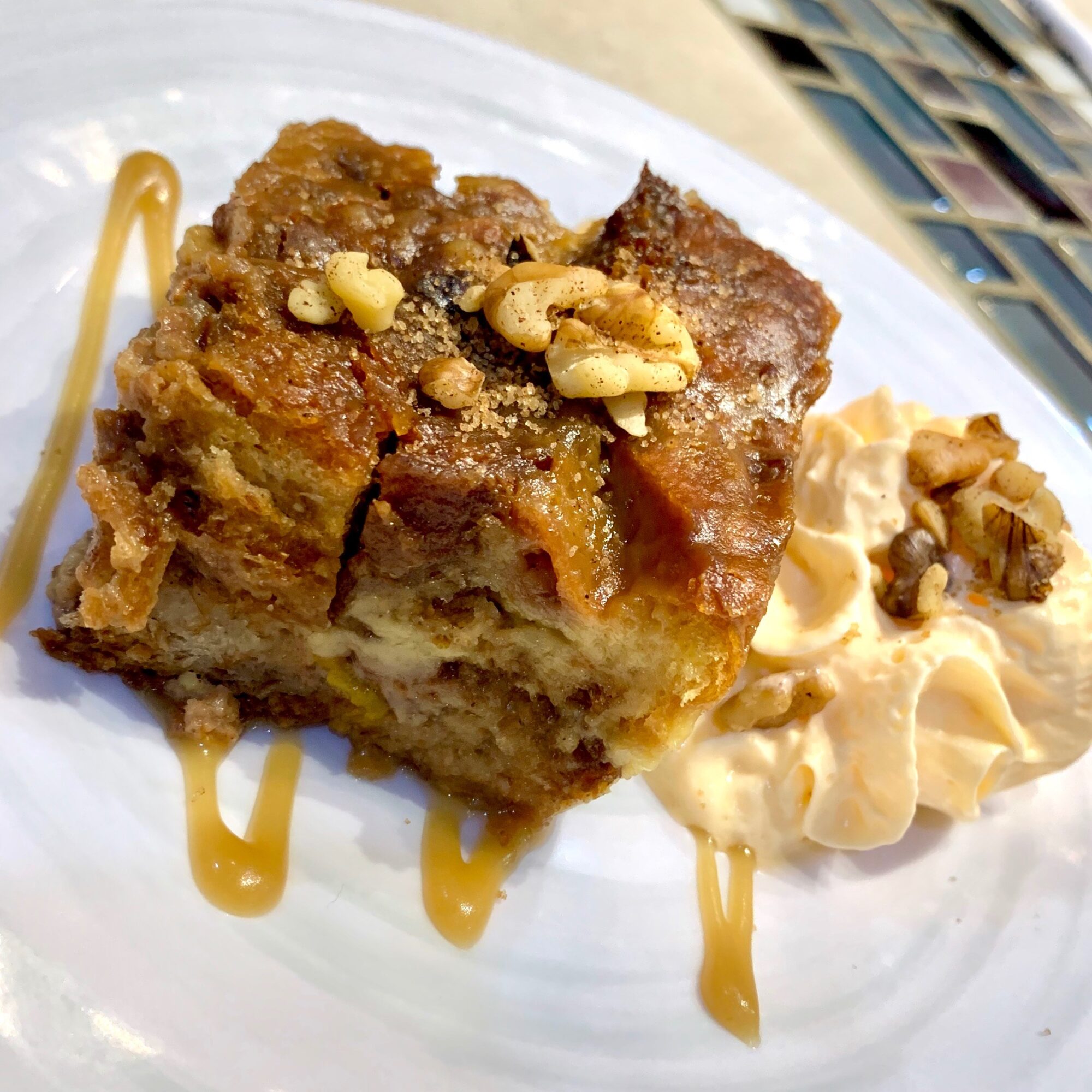 A slice of dessert with nuts, caramel drizzle, and a side of whipped cream with nuts on a white plate.