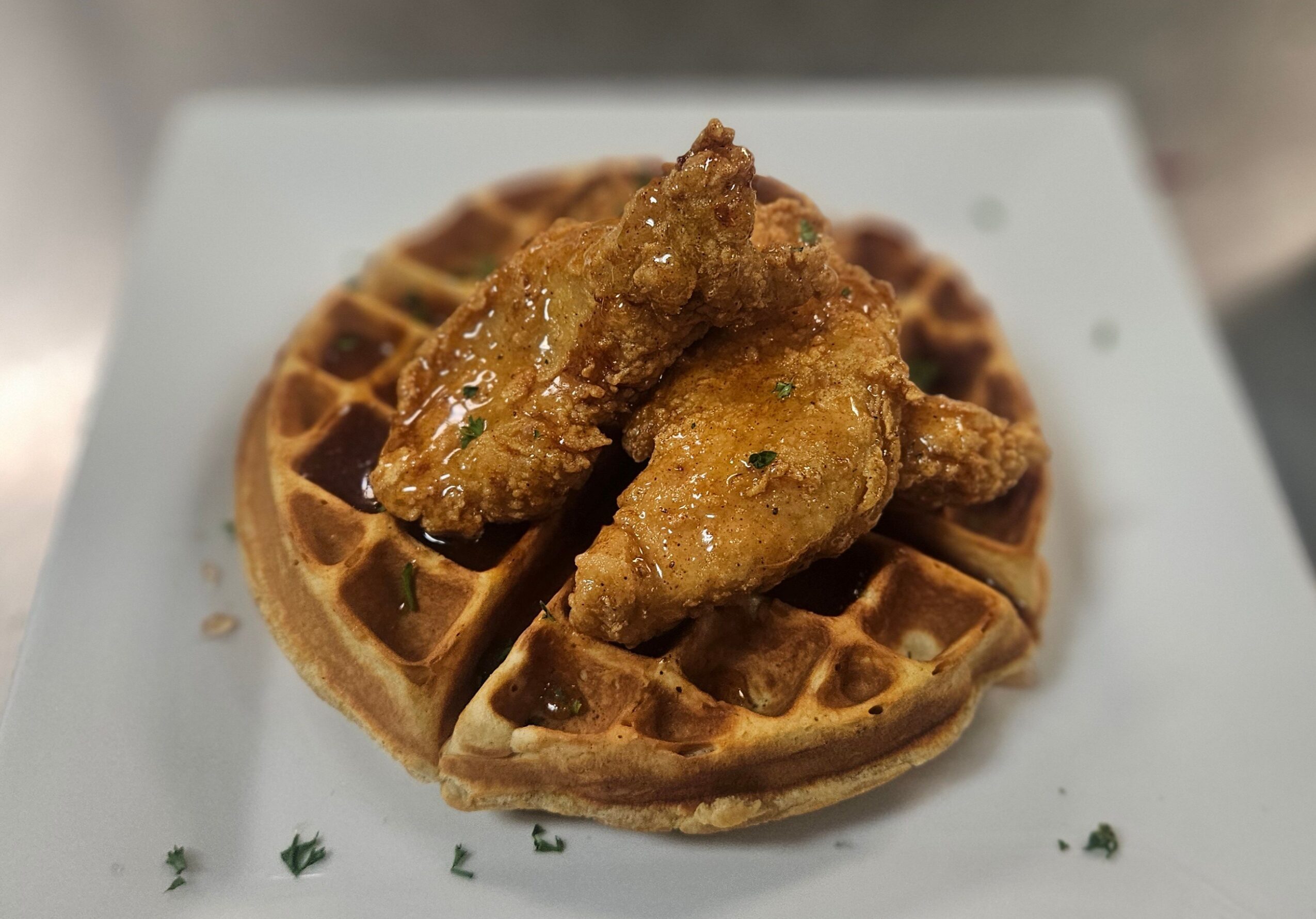 Waffle topped with fried chicken pieces on a white plate, garnished with herbs, close-up view.