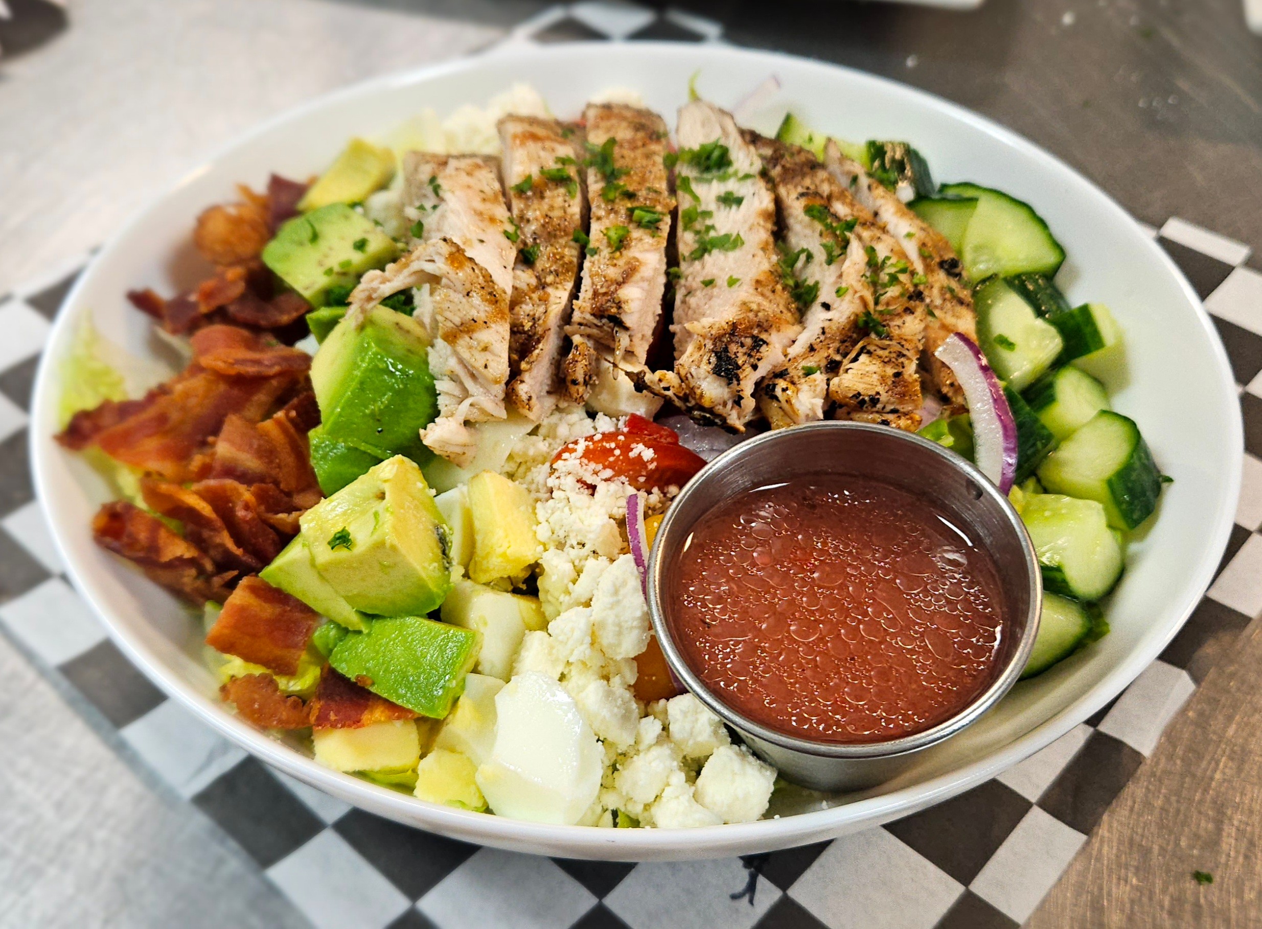 Bowl with grilled chicken, cucumber, avocado, tomato, onion, crumbled cheese, and a small cup of sauce on a checkered paper