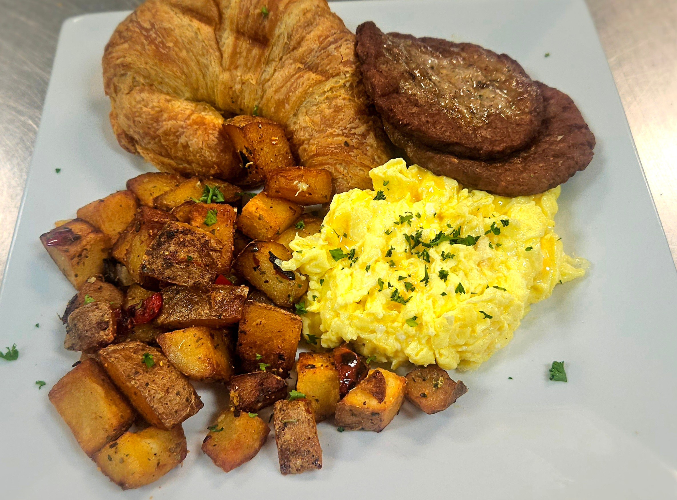 Croissant, steak, scrambled eggs, and roasted potatoes on a white plate.