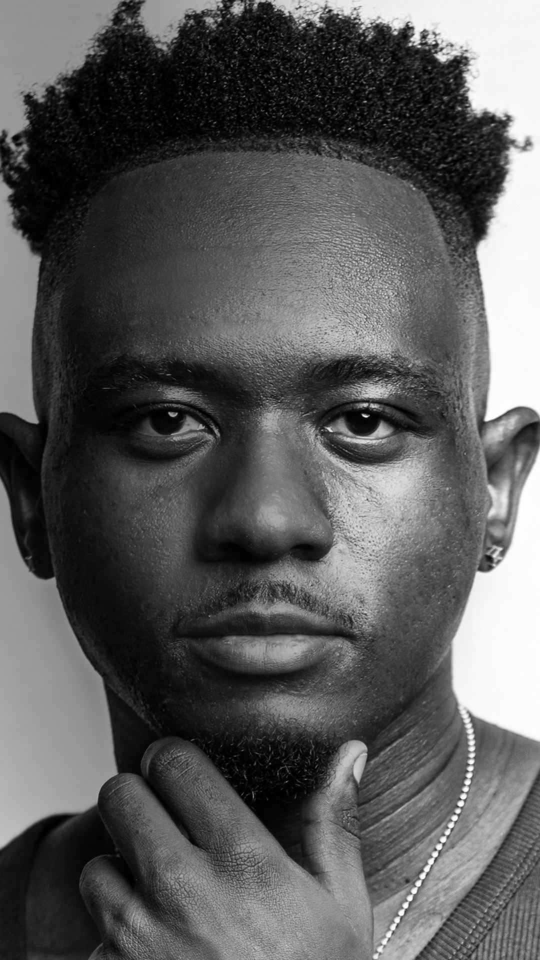 Close-up of a young man with short curly hair, looking directly at the camera, hand near chin, in black and white.