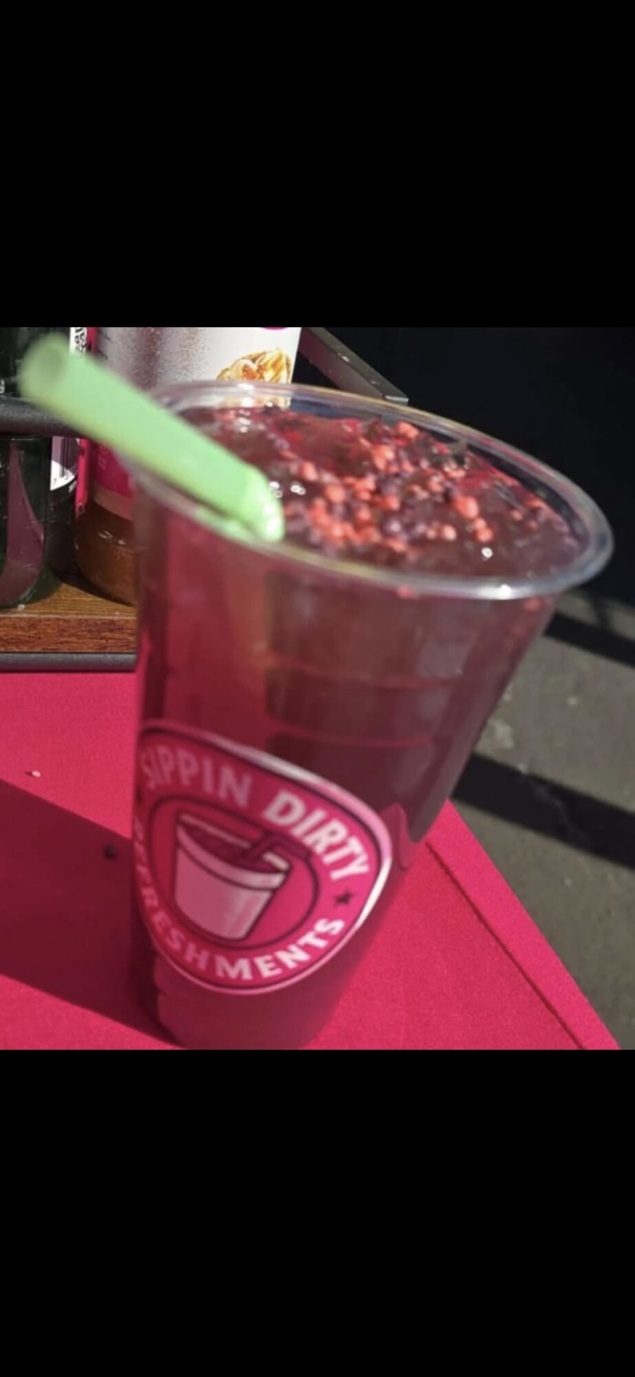 Pink cup with a green straw, topped with a dark beverage and sprinkles, on a pink surface.