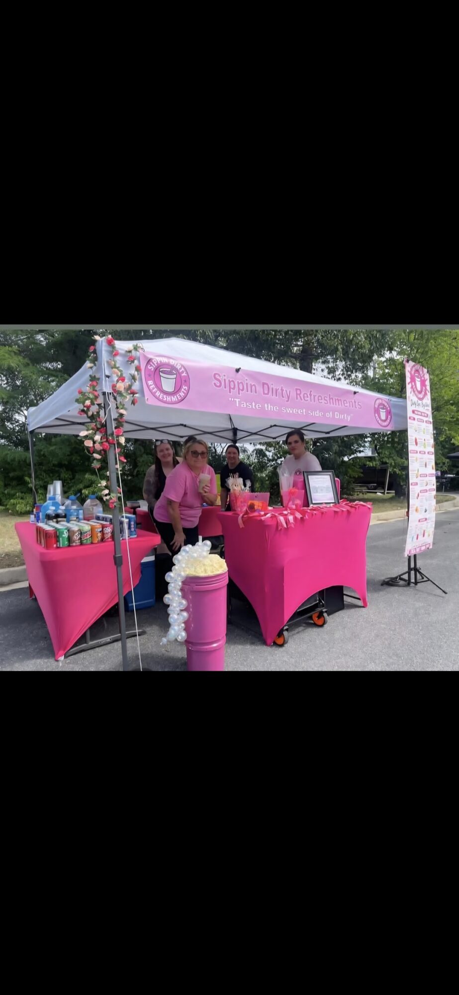 Pink canopy tent with three people behind a table, outdoor setting with trees in background.
