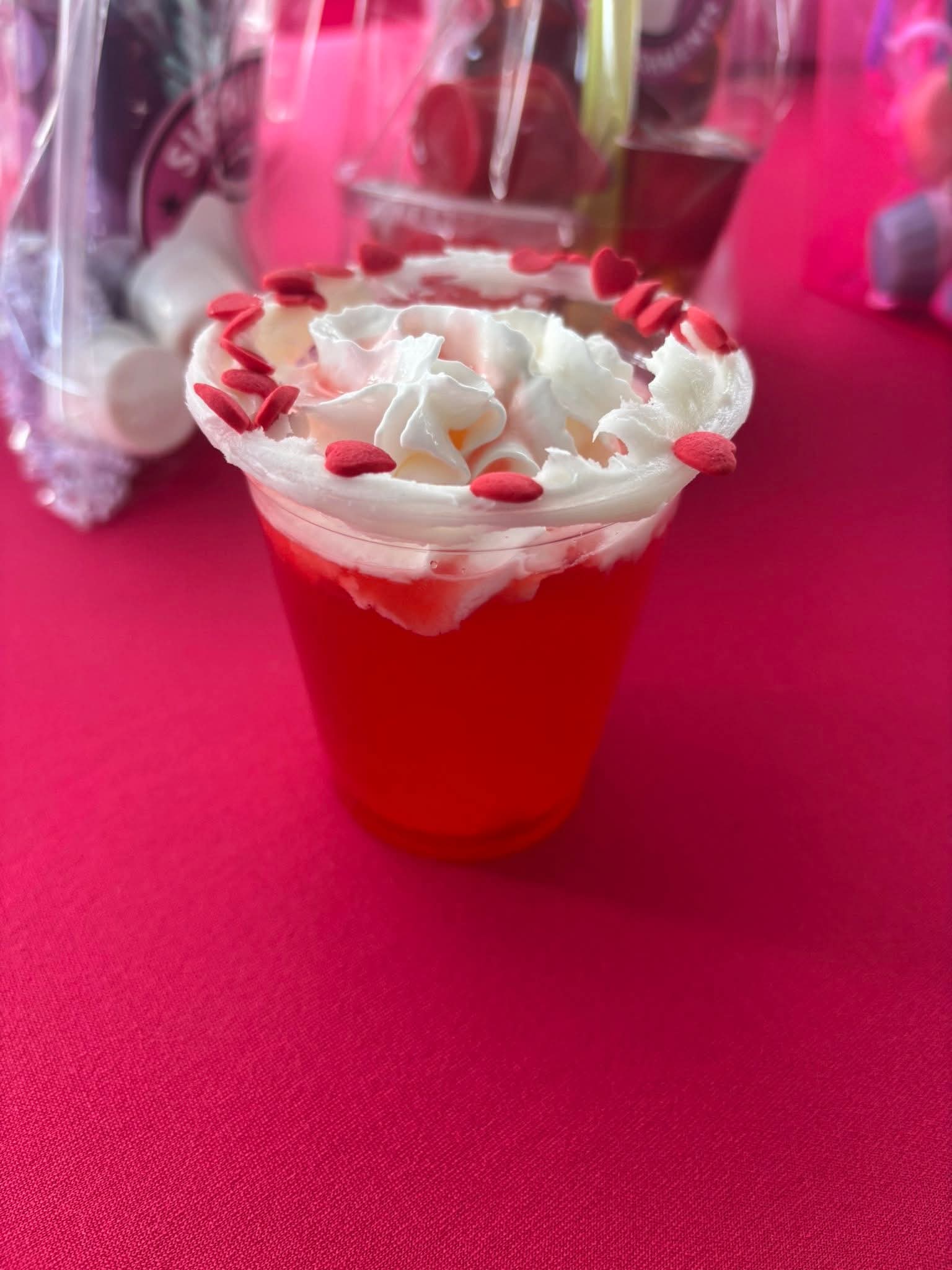 Red and white decorated dessert cup with whipped cream and heart-shaped sprinkles on a pink surface.