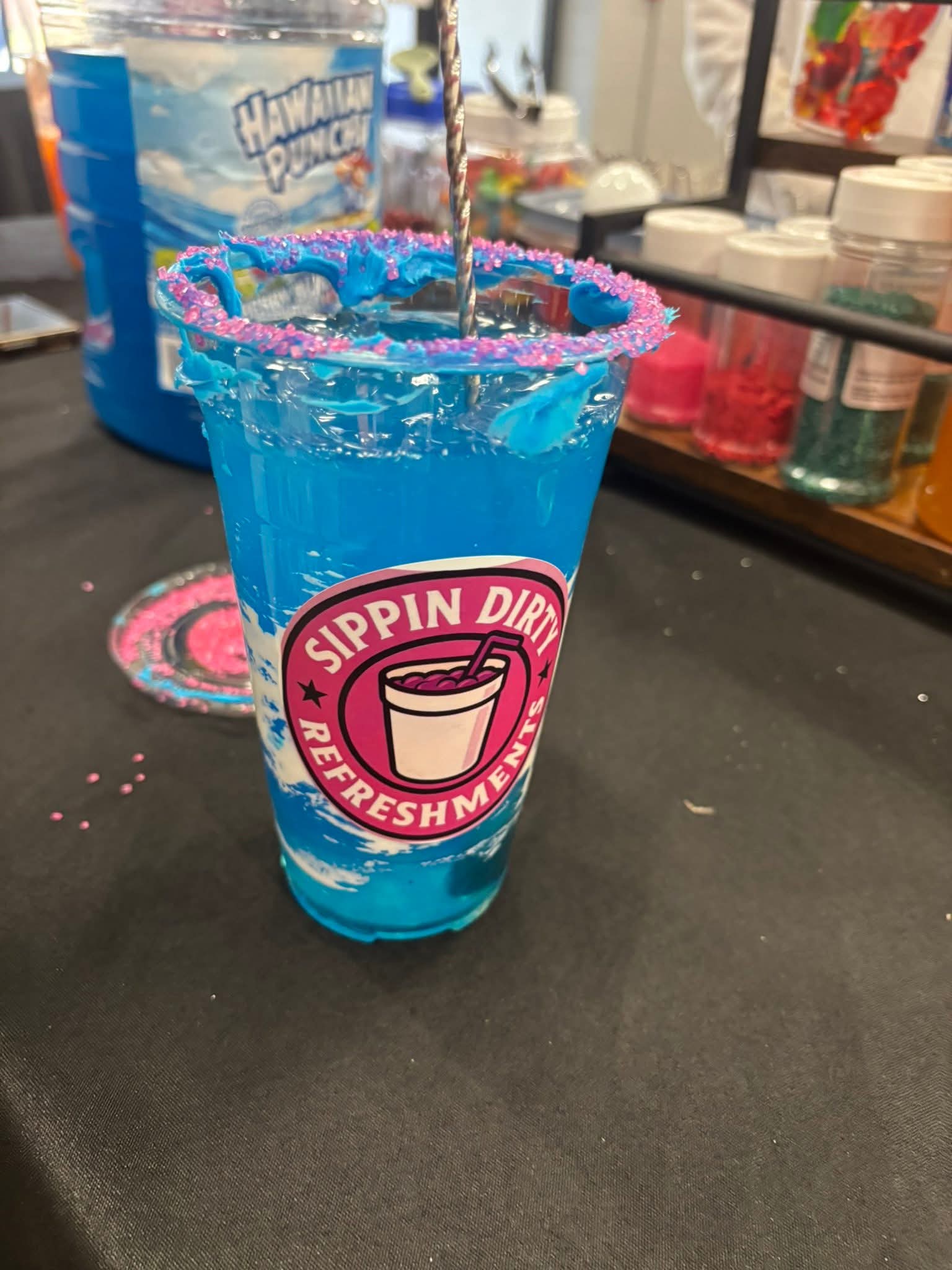 Blue beverage in a clear cup with a pink and white logo, on a black surface with store items in background.