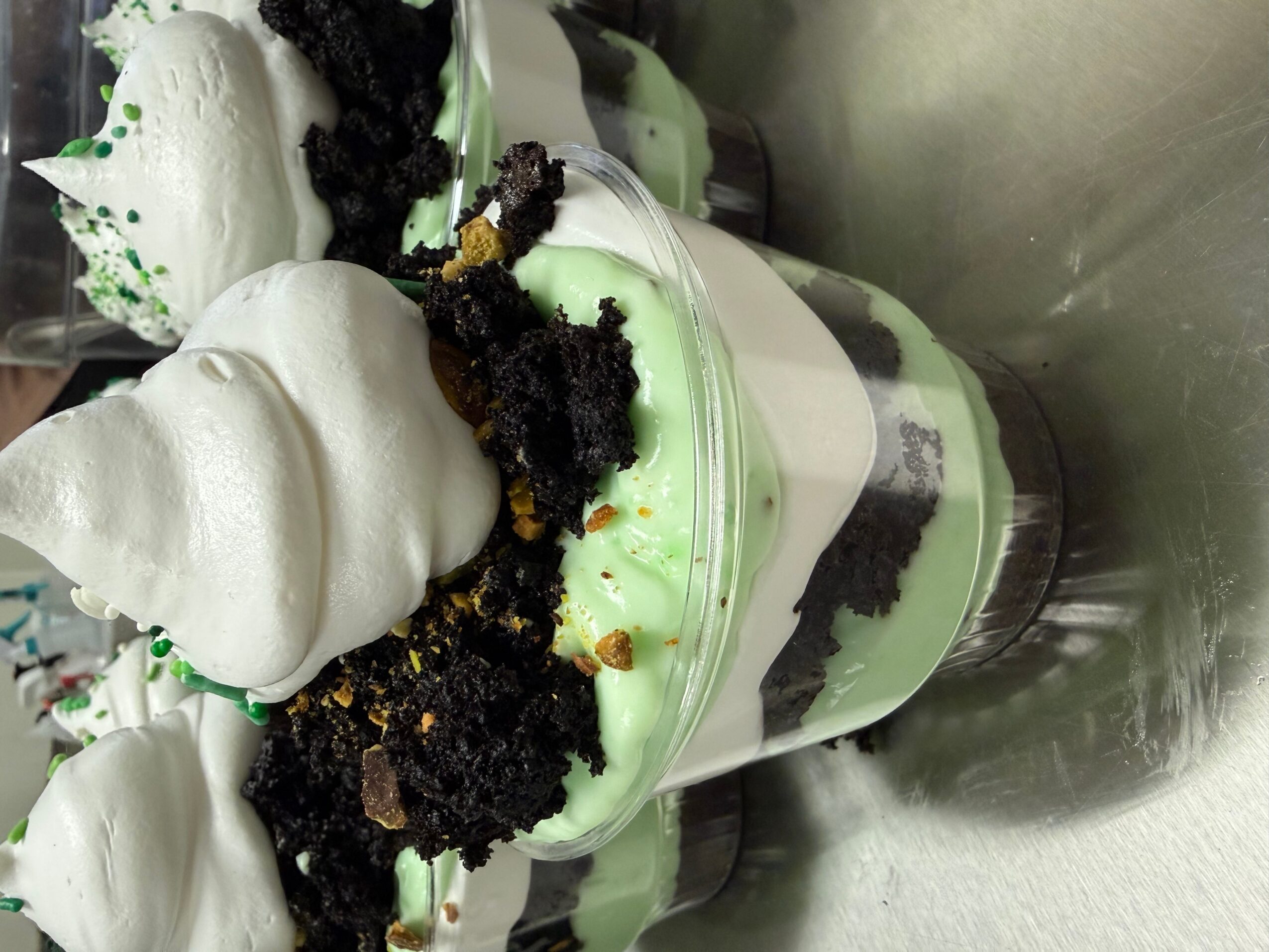 A layered dessert with green and white cream, topped with crumbled cookies and meringue, in a clear glass cup.