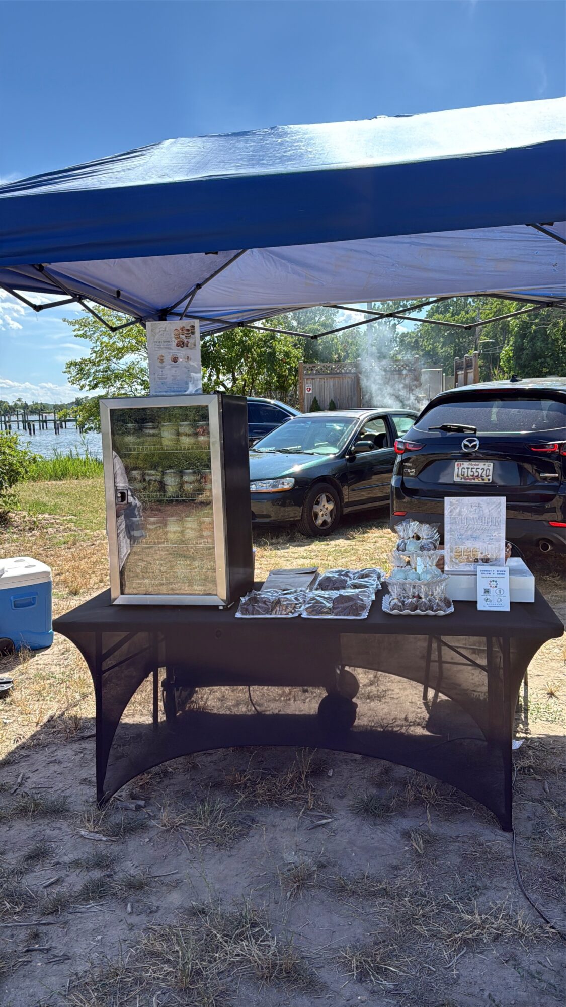 Outdoor table with a glass display case and items for sale, under a canopy, with parked cars and trees in background.
