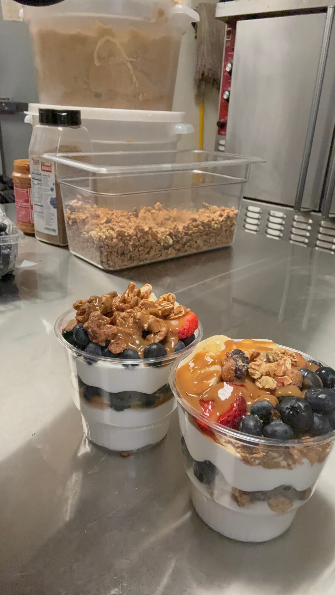 Two dessert cups with layered ingredients, topped with nuts, fruits, and granola, on a reflective surface in a kitchen.