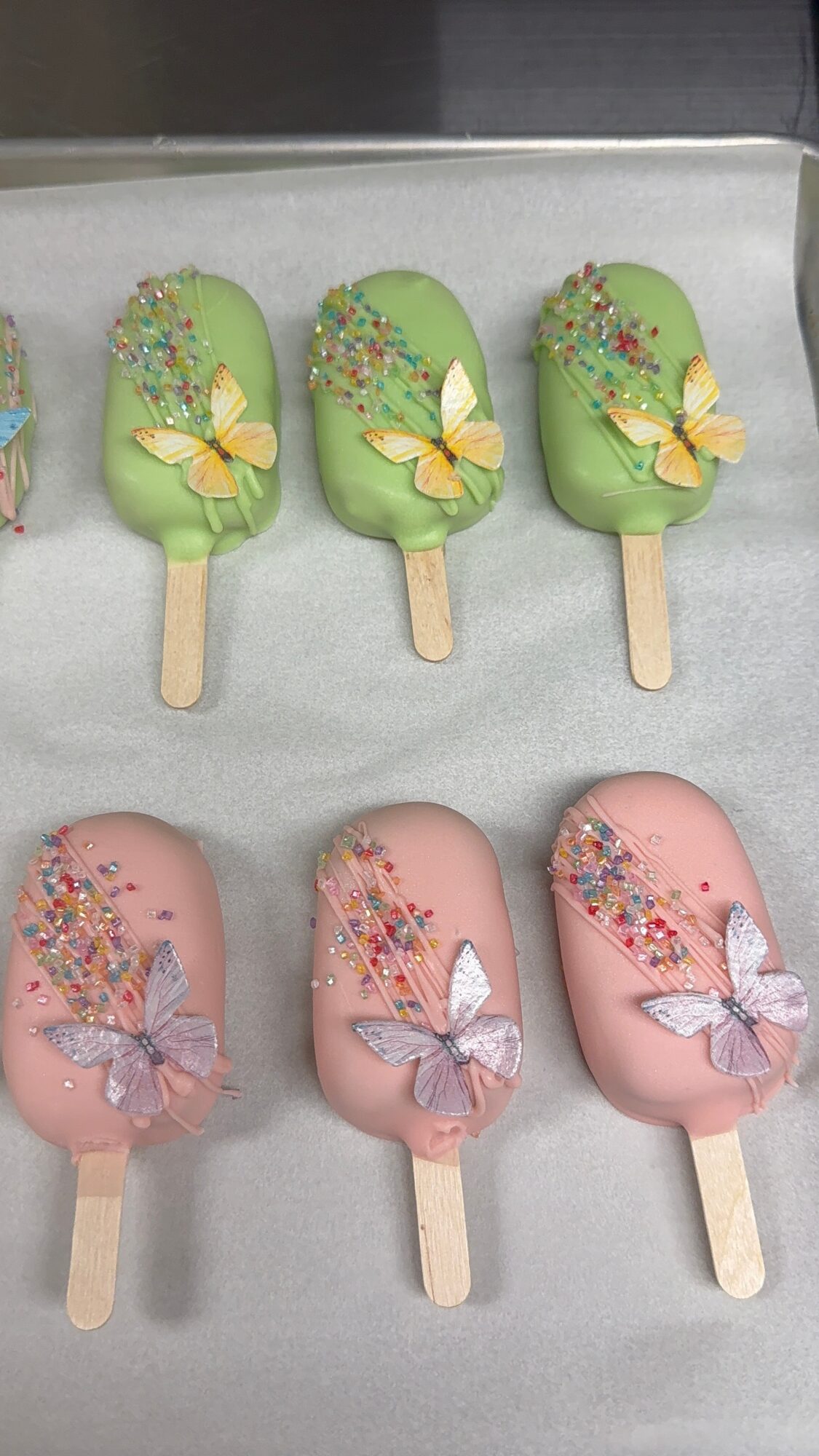 Six decorated ice cream bars with butterfly designs, three green and three pink, on a white surface.