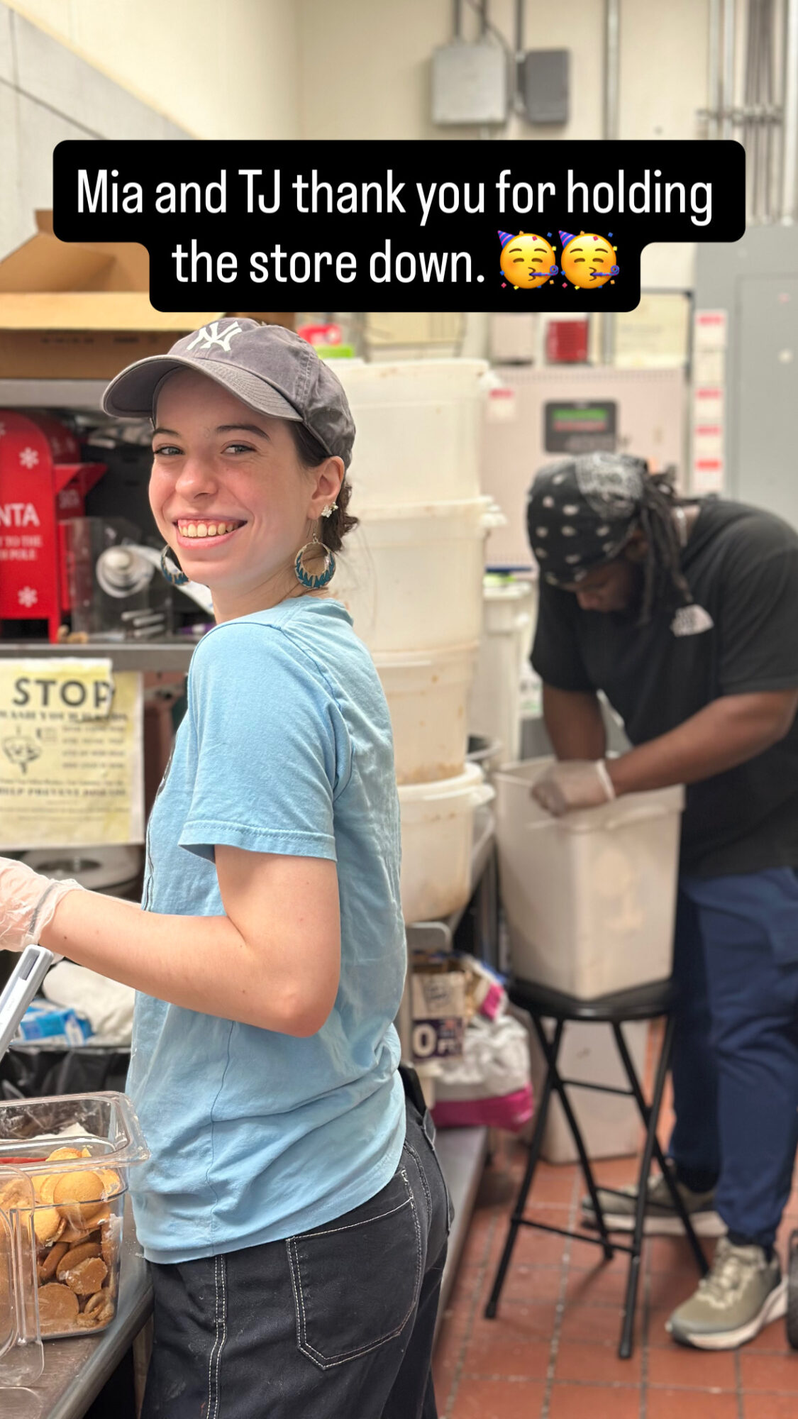 Young woman smiling in a store, holding a bag, with a person working behind her. Store shelves and a sign are visible.