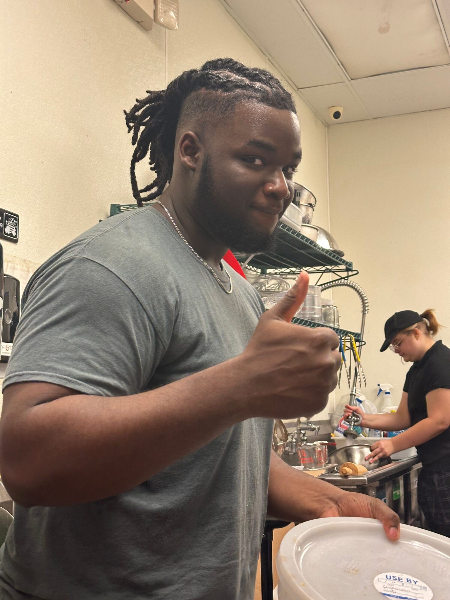 Person with braided hair and gray shirt pointing with finger in a kitchen or dining area.