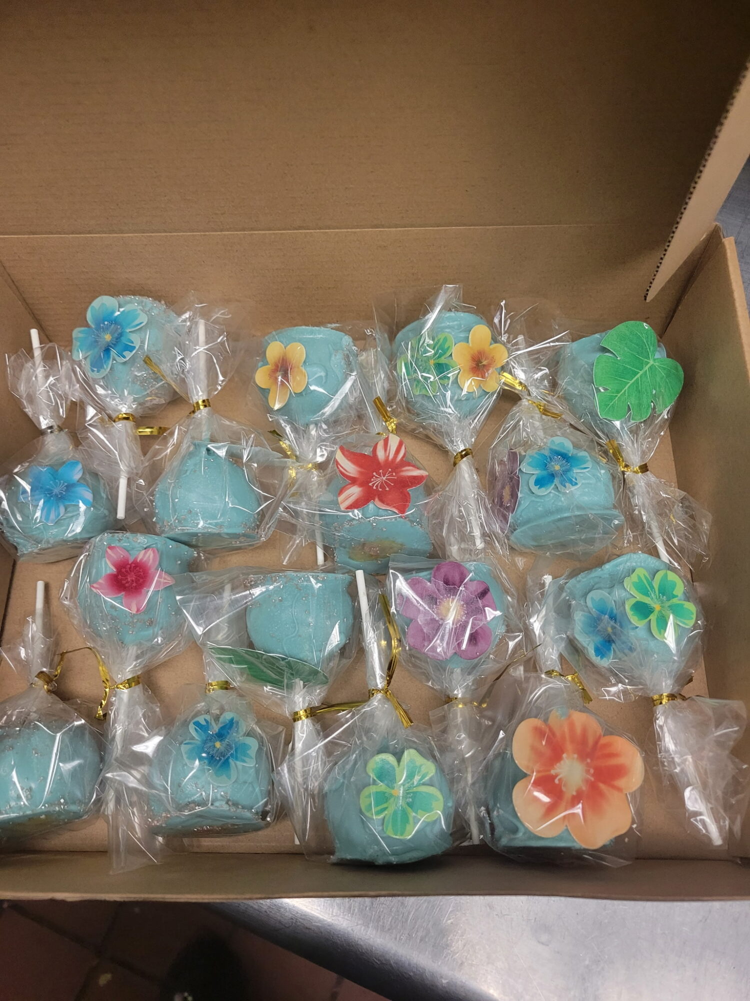 Box of decorated cookies with colorful flower and leaf designs, individually wrapped in clear plastic, on a brown surface.
